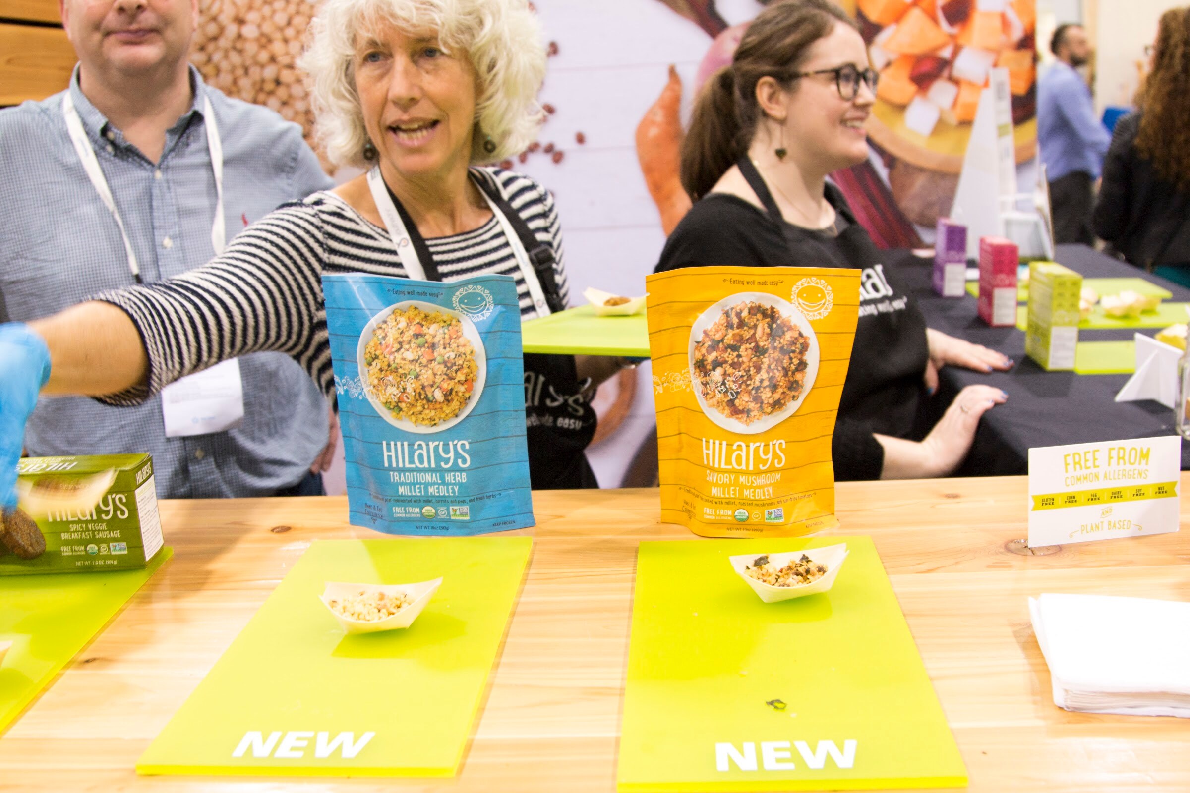 Counter with pouches of Hilary's Traditional Herb and Savory Mushroom millet medley, small sample bowls and staff members in aprons smiling behind.