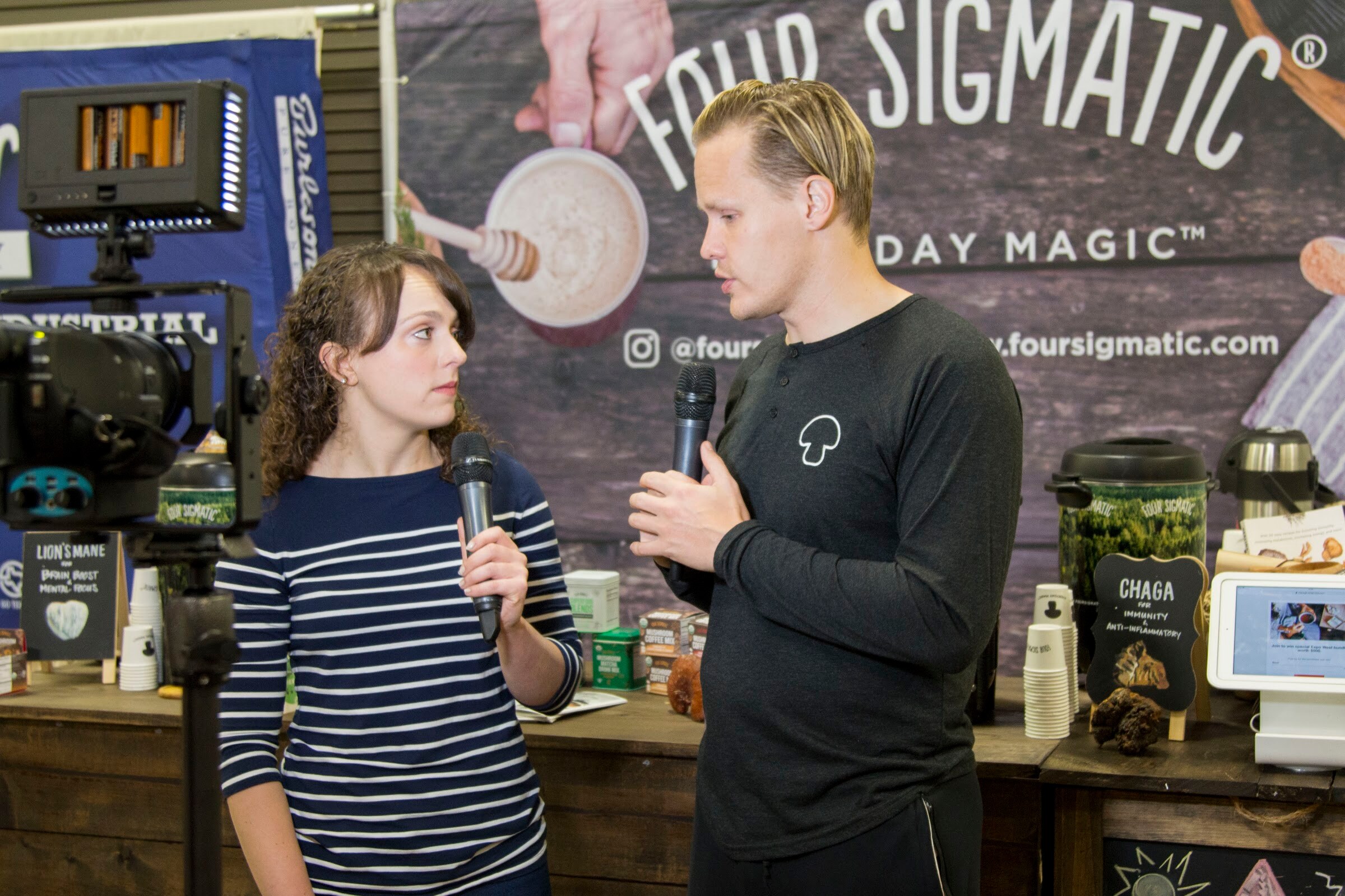 Woman with a microphone and a man in a dark shirt with a mushroom logo standing in front of a Four Sigmatic backdrop, with a video light and camera at left.