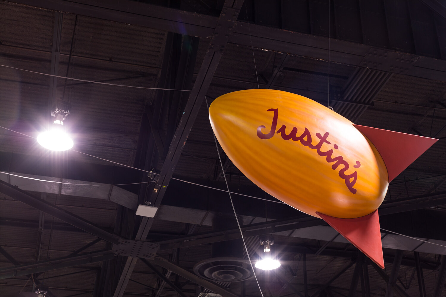 Orange blimp-shaped sign with the word Justin's hanging from a ceiling with lights and ductwork.
