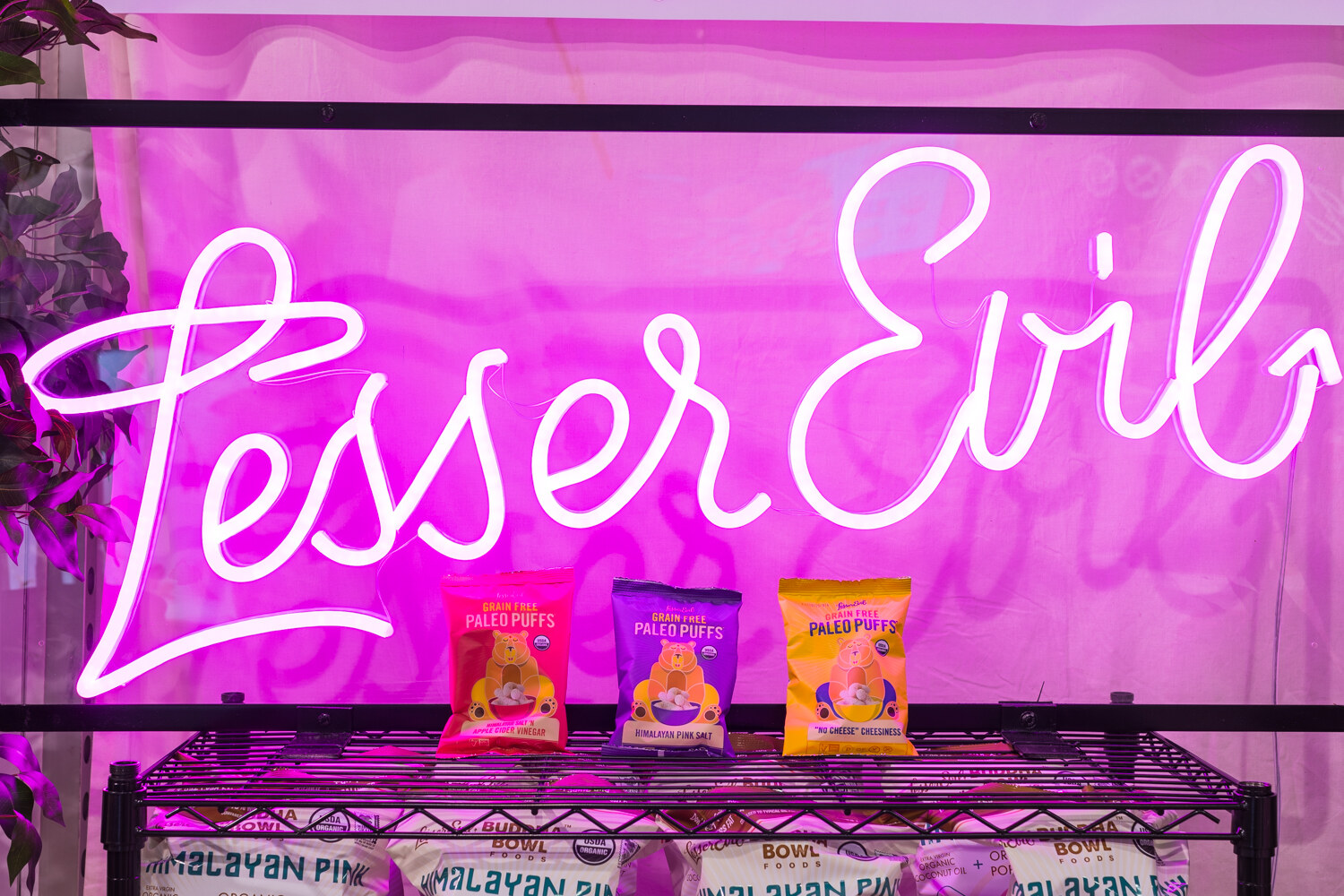 Pink neon script sign reading Lesser Evil above a wire shelf with three small bags of paleo puffs.
