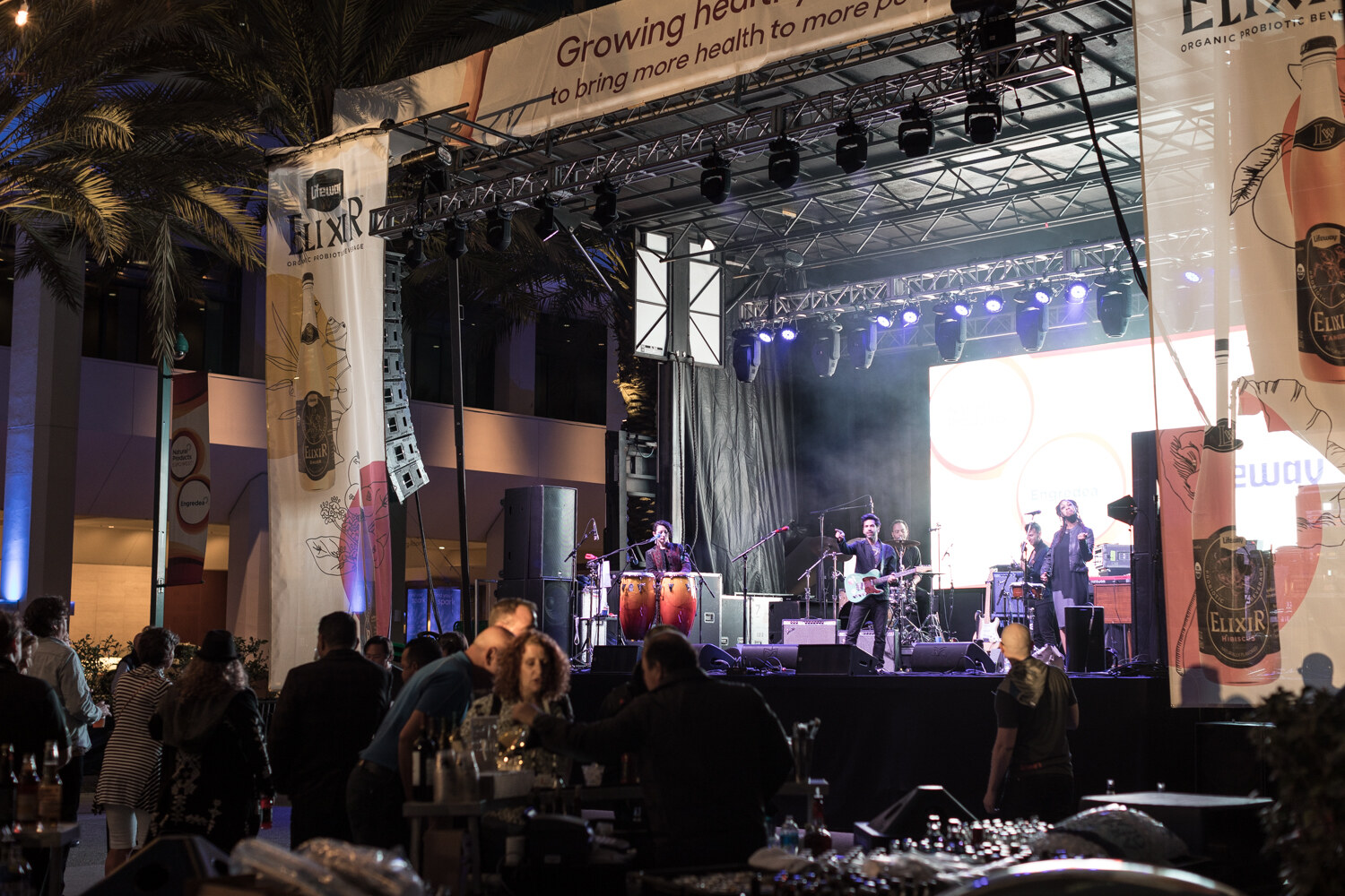 Dusk scene of a band performing on a stage with truss lighting and tall banners, with a crowd in the foreground.