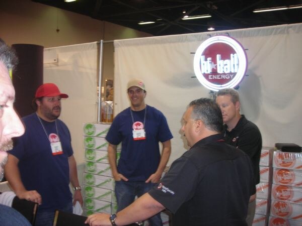 Several men wearing badges stand in a booth under a lit hi-ball Energy sign, with stacked cases of product on both sides; one man in the foreground faces away from the camera.