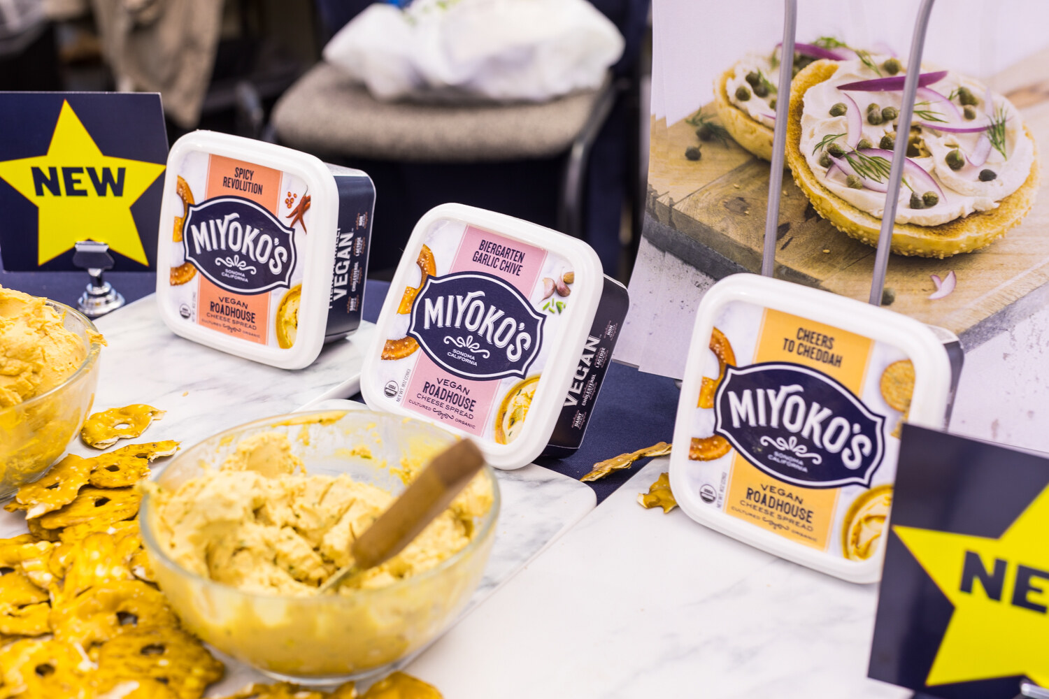 Tubs of Miyoko's vegan cream cheese spreads next to a bowl of spread and a bagel topped with cream cheese, with a yellow star reading NEW.