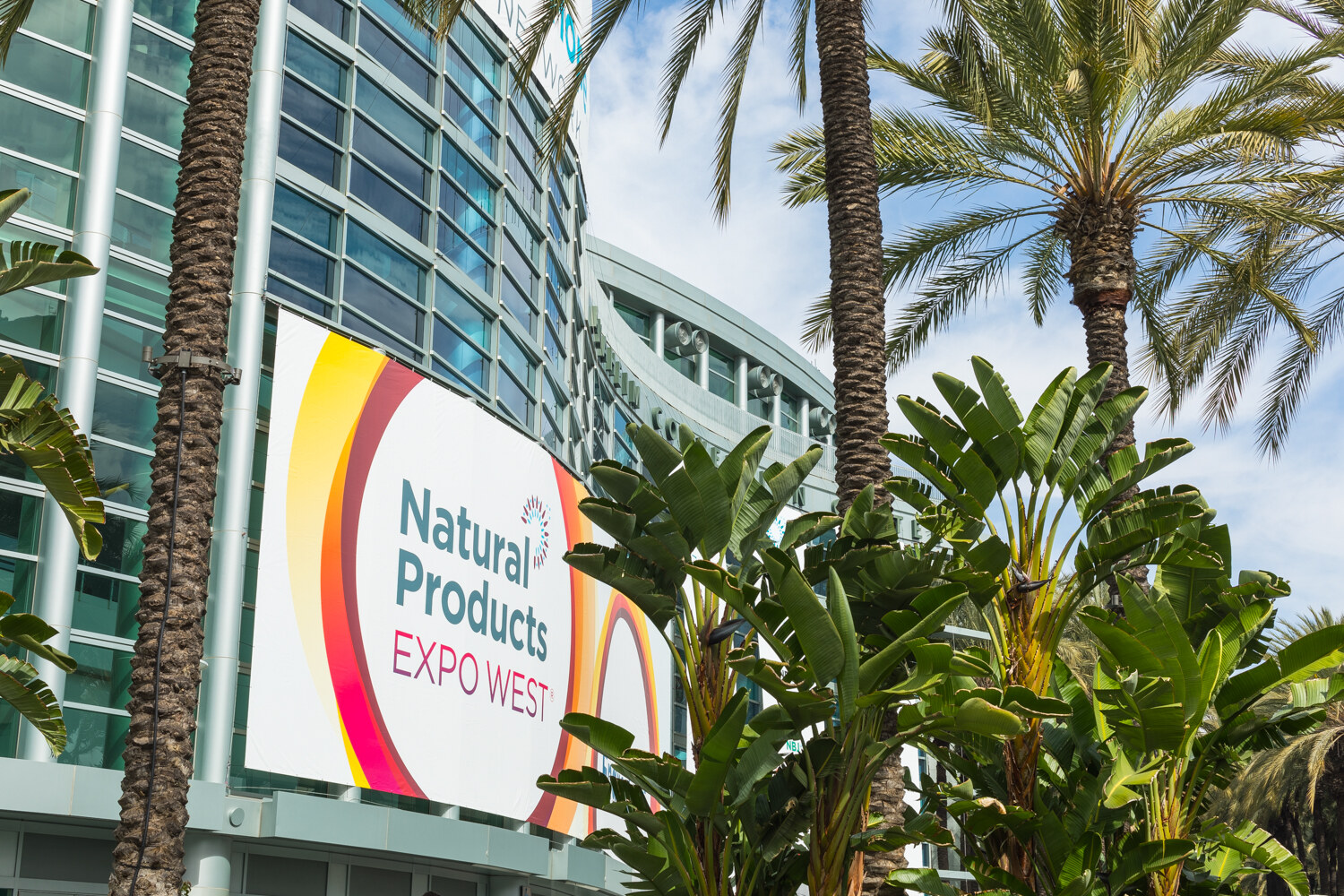 Large Natural Products Expo West sign on the glass facade of a convention center, framed by palm trees and tropical plants under a blue sky.