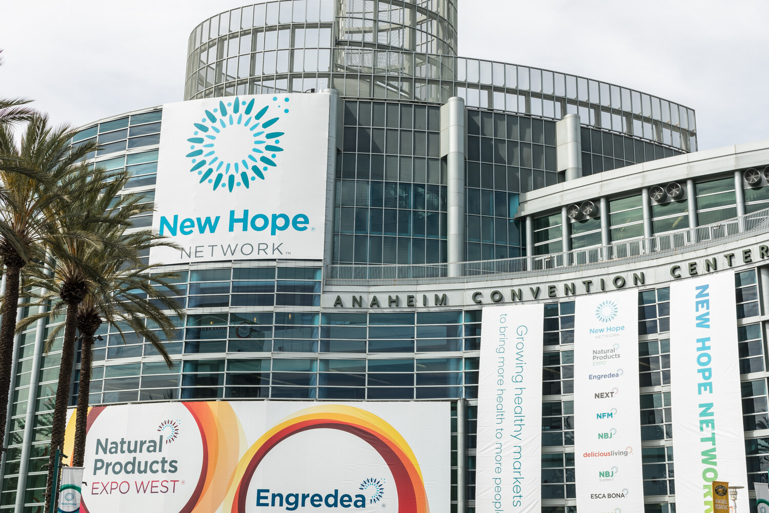 Convention center facade with a large New Hope Network banner, Natural Products Expo West and Engredea signs, and tall vertical banners beside palm trees.