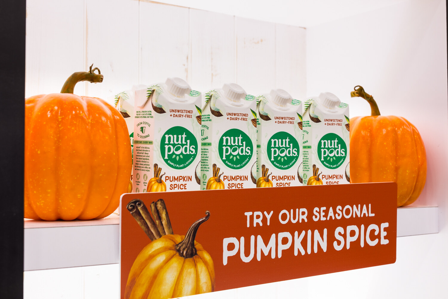 Four nutpods cartons flanked by orange pumpkins with a sign reading 'Try our seasonal Pumpkin Spice'.