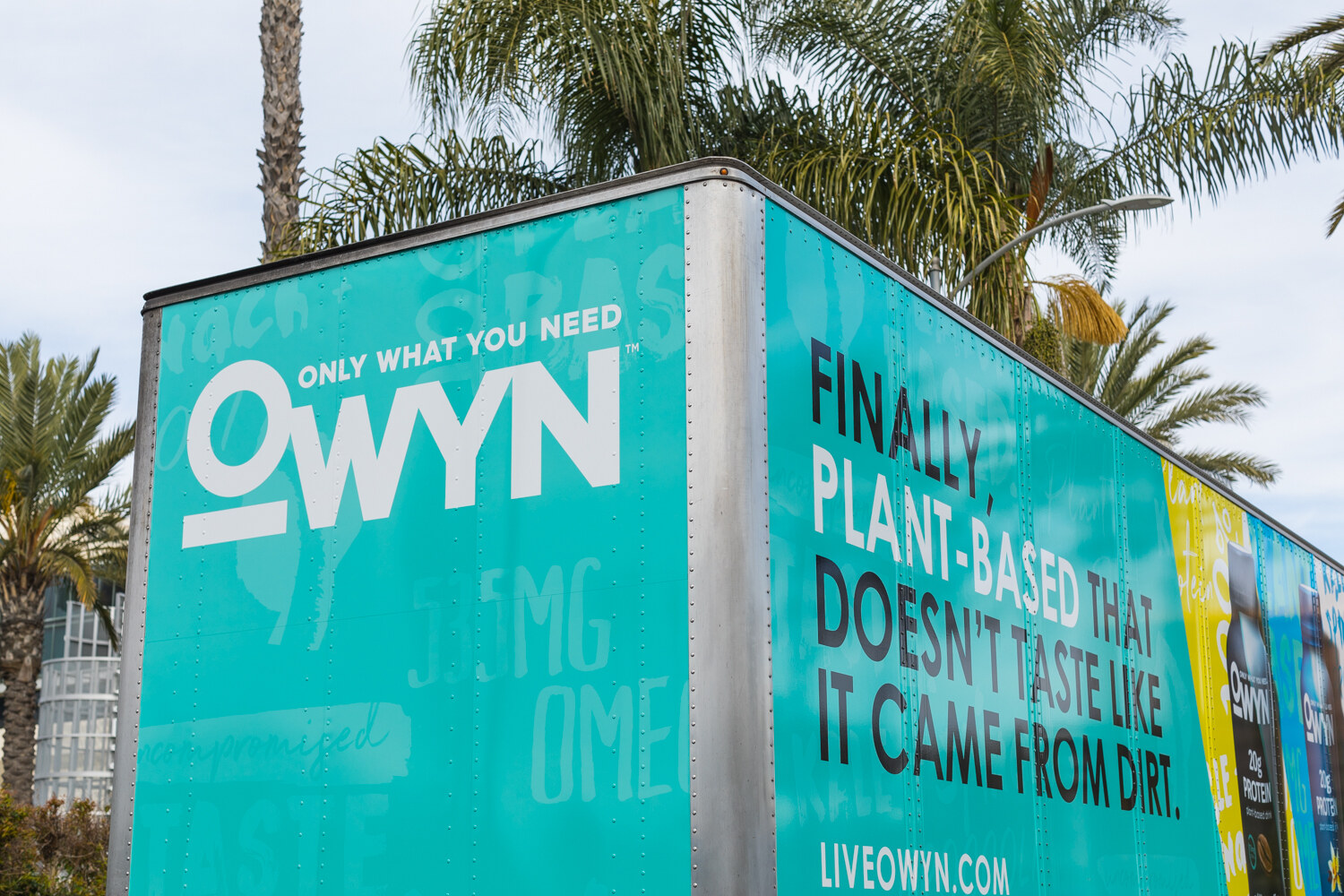 Teal truck wrapped with the OWYN logo and the slogan 'Finally, plant-based that doesn't taste like it came from dirt' with palm trees behind it.