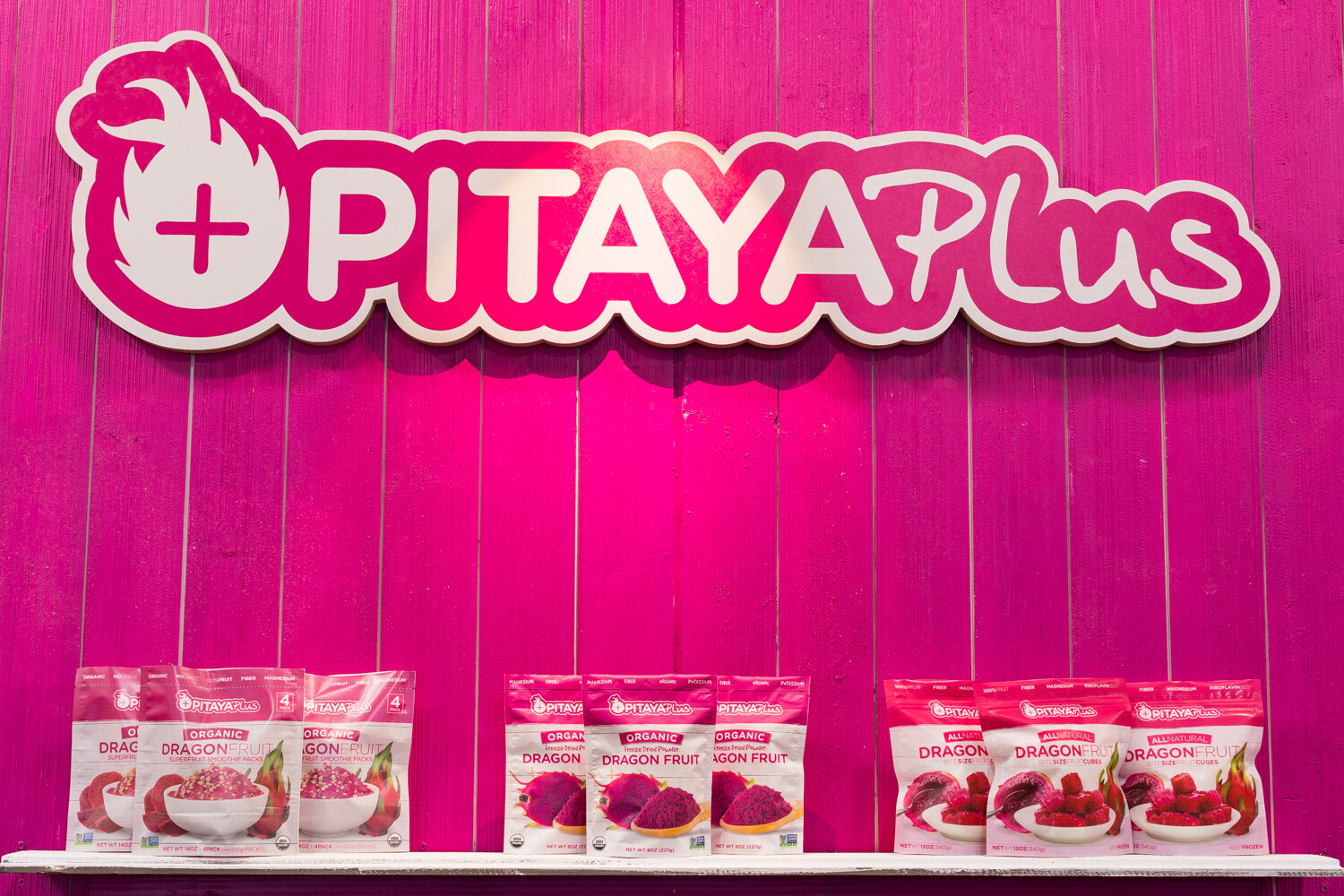 Pink wall with a large Pitaya Plus sign and three shelves of small pink dragon fruit packets below.