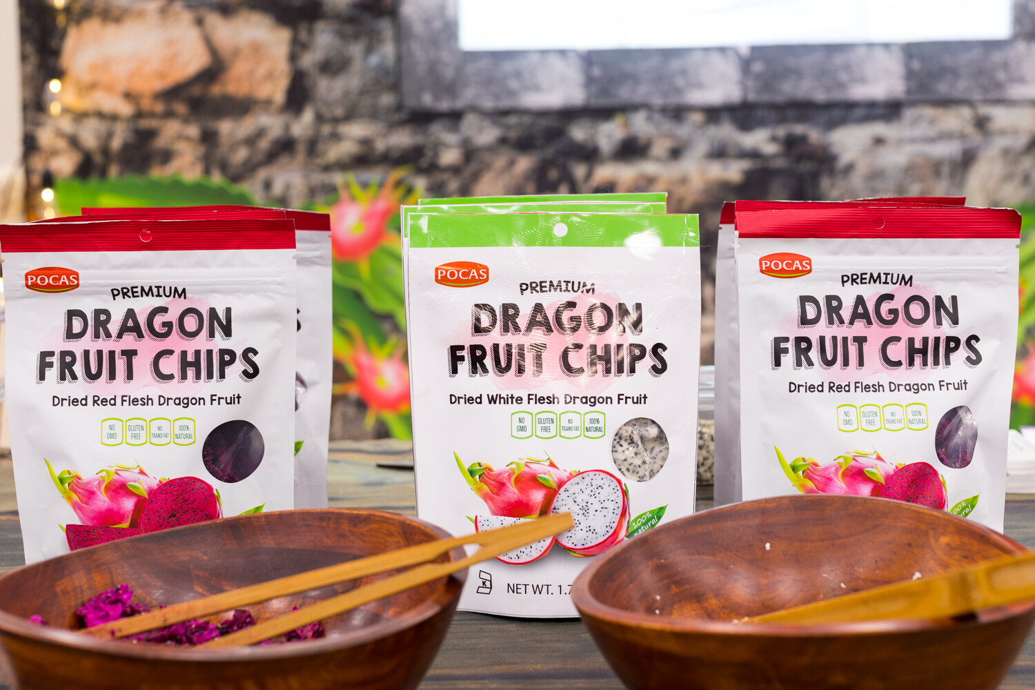 Three bags of Pocas premium dragon fruit chips on a table with two wooden bowls, with dragon fruit art in the background.