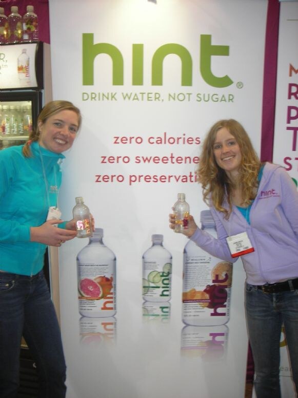 Two smiling women hold small bottles of hint water in front of a hint banner reading "Drink water, not sugar," with bottle graphics below and a refrigerated display at left.