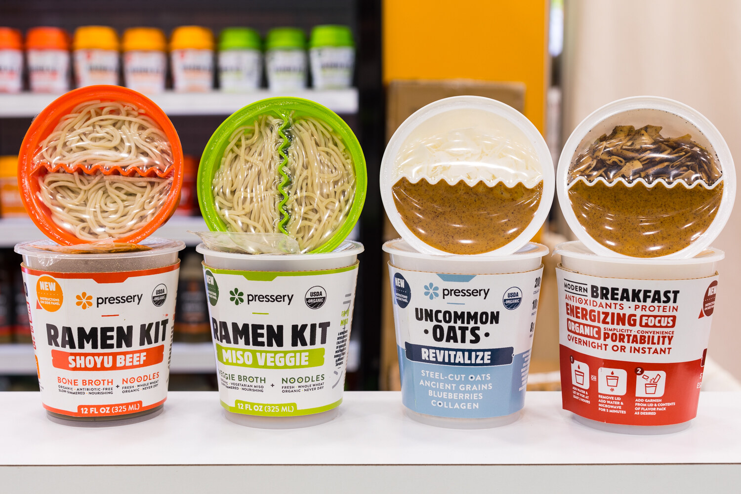 Four Pressery cups: two ramen kits with noodles and broth, and two oat cups, on a white shelf.