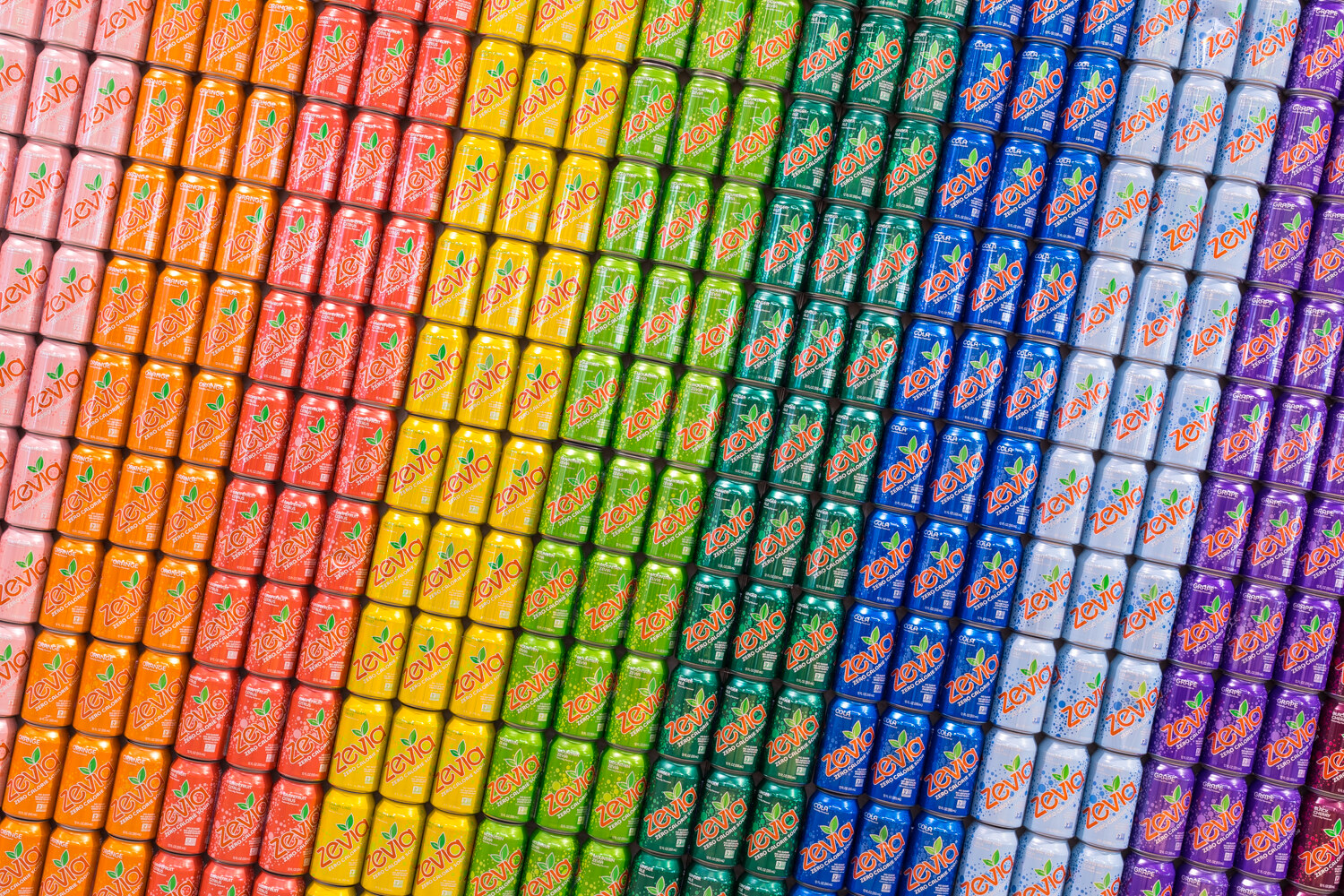 Close-up of rows of Zevia soda cans arranged in a diagonal color gradient from orange through green and blue.