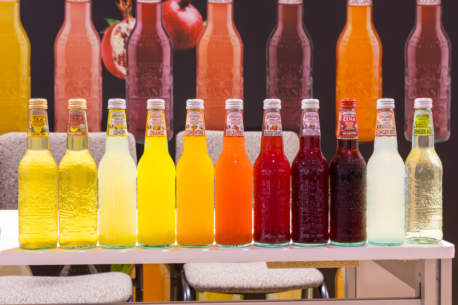 Eleven glass bottles of organic soda in colors from yellow to dark red standing on a white shelf.