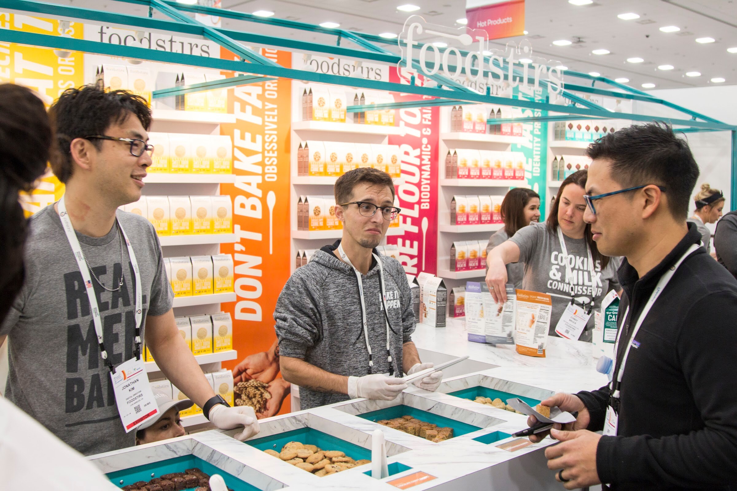 Busy booth with a teal frame and a foodstirs logo, three staff in the center serving samples and a visitor in black leaning in from the right.