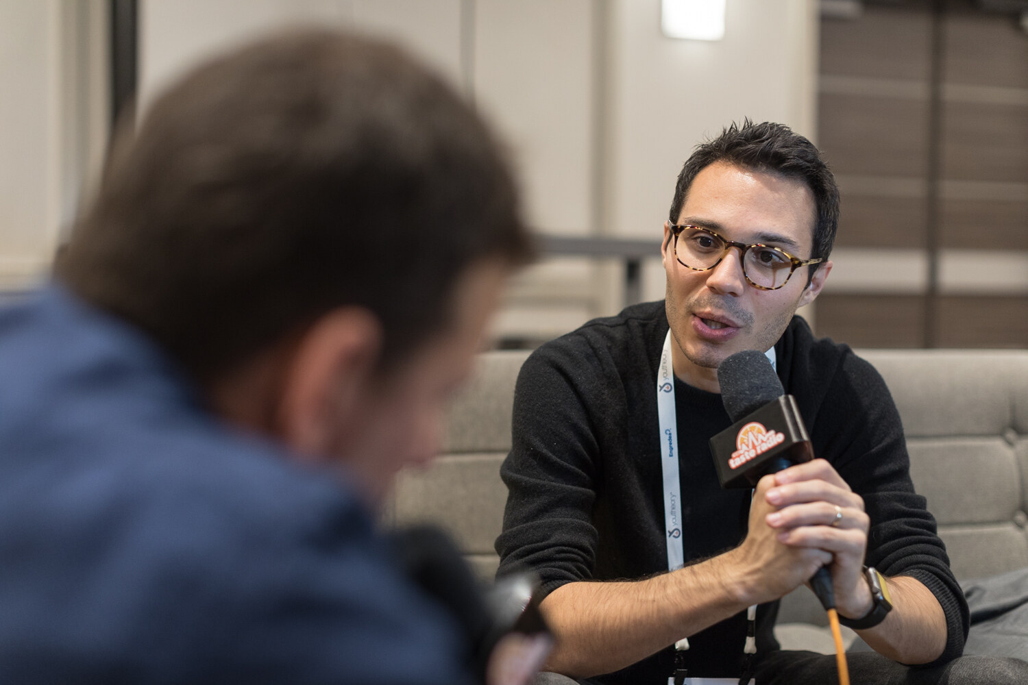 Man with glasses and a lanyard speaking into a microphone, with the blurred back of another person's head in the left foreground.