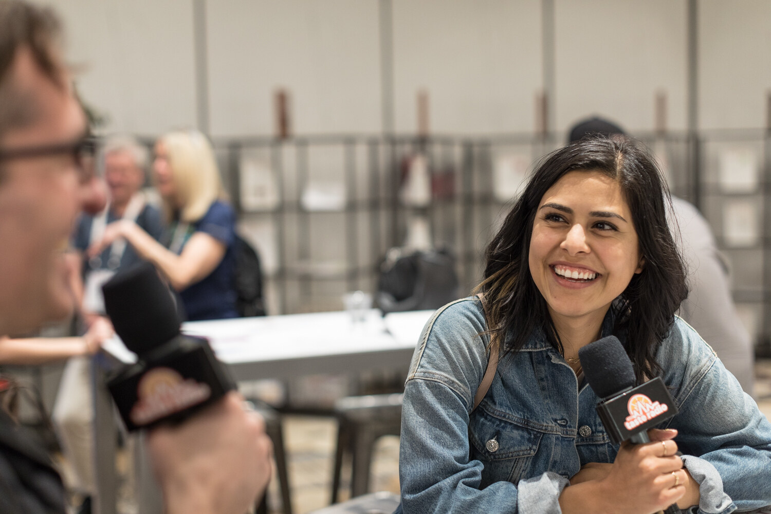 Woman in a denim jacket smiling broadly and holding a microphone with a Taste Radio flag while a man with glasses sits in the foreground.