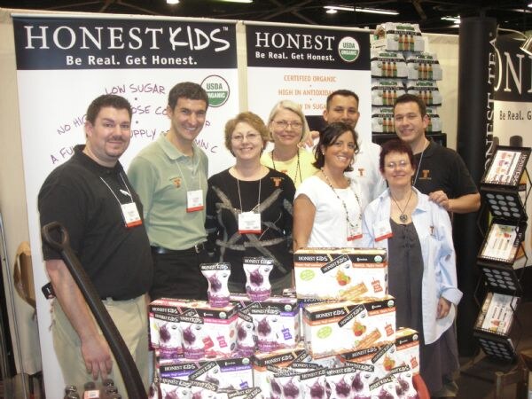 Seven people in a group pose behind stacked boxes of Honest Kids drinks, in front of Honest Kids banners reading "Be Real. Get Honest." and a USDA organic seal.