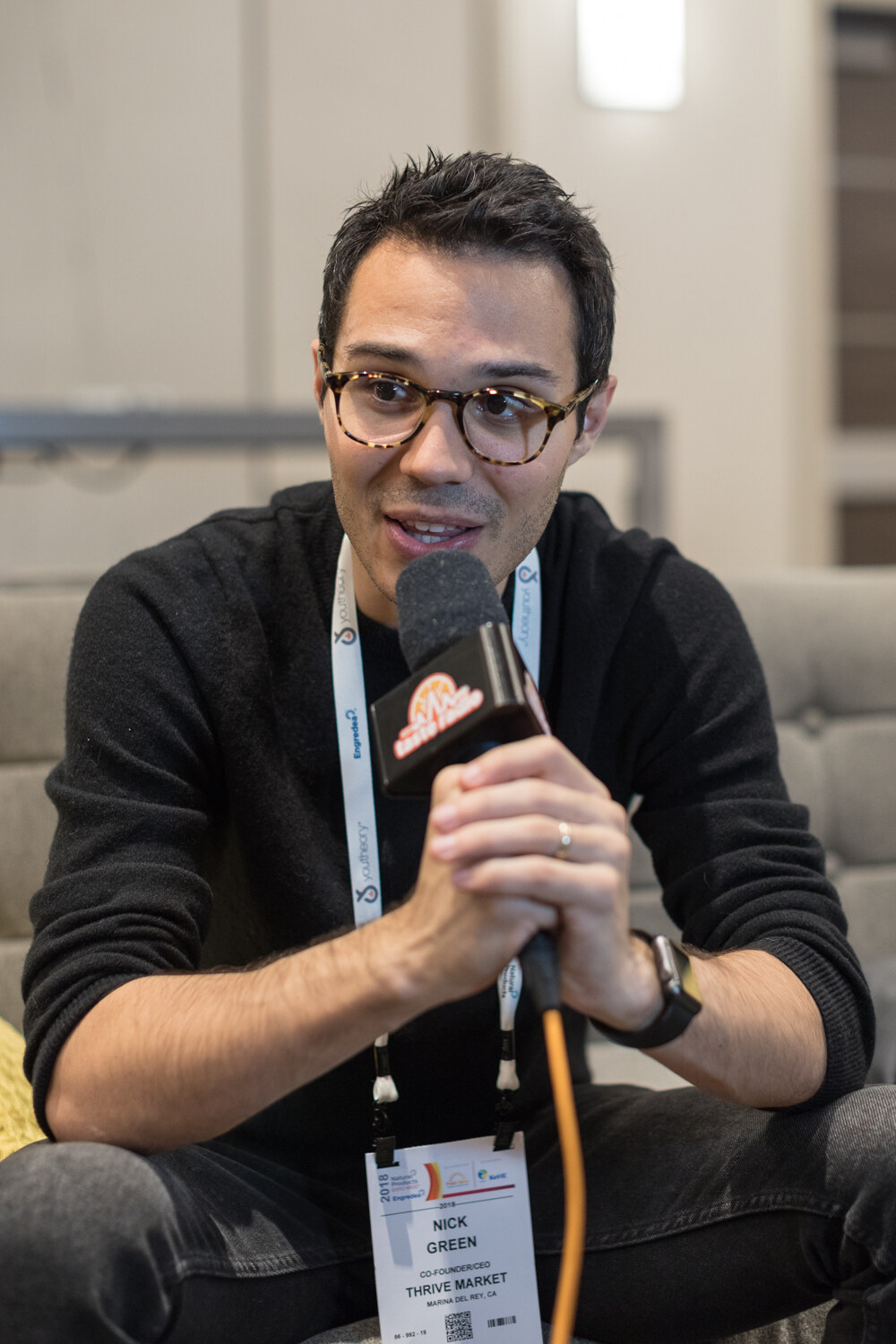 Man with round glasses and a lanyard holding a Taste Radio microphone with an orange cable, seated on a grey couch.