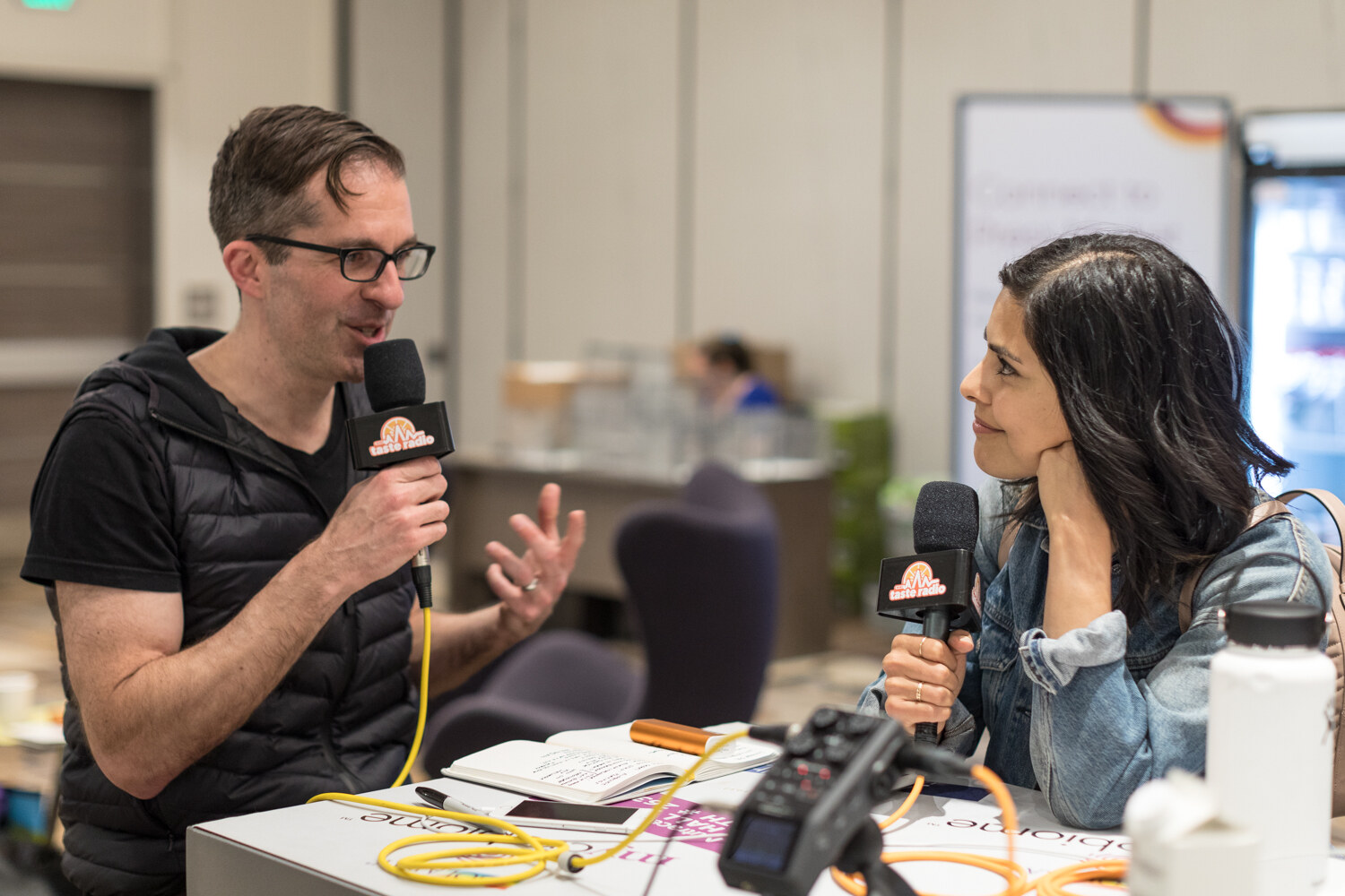 Two people seated at a table with microphones carrying Taste Radio flags; the man in a black vest gestures while a woman in a denim jacket listens.