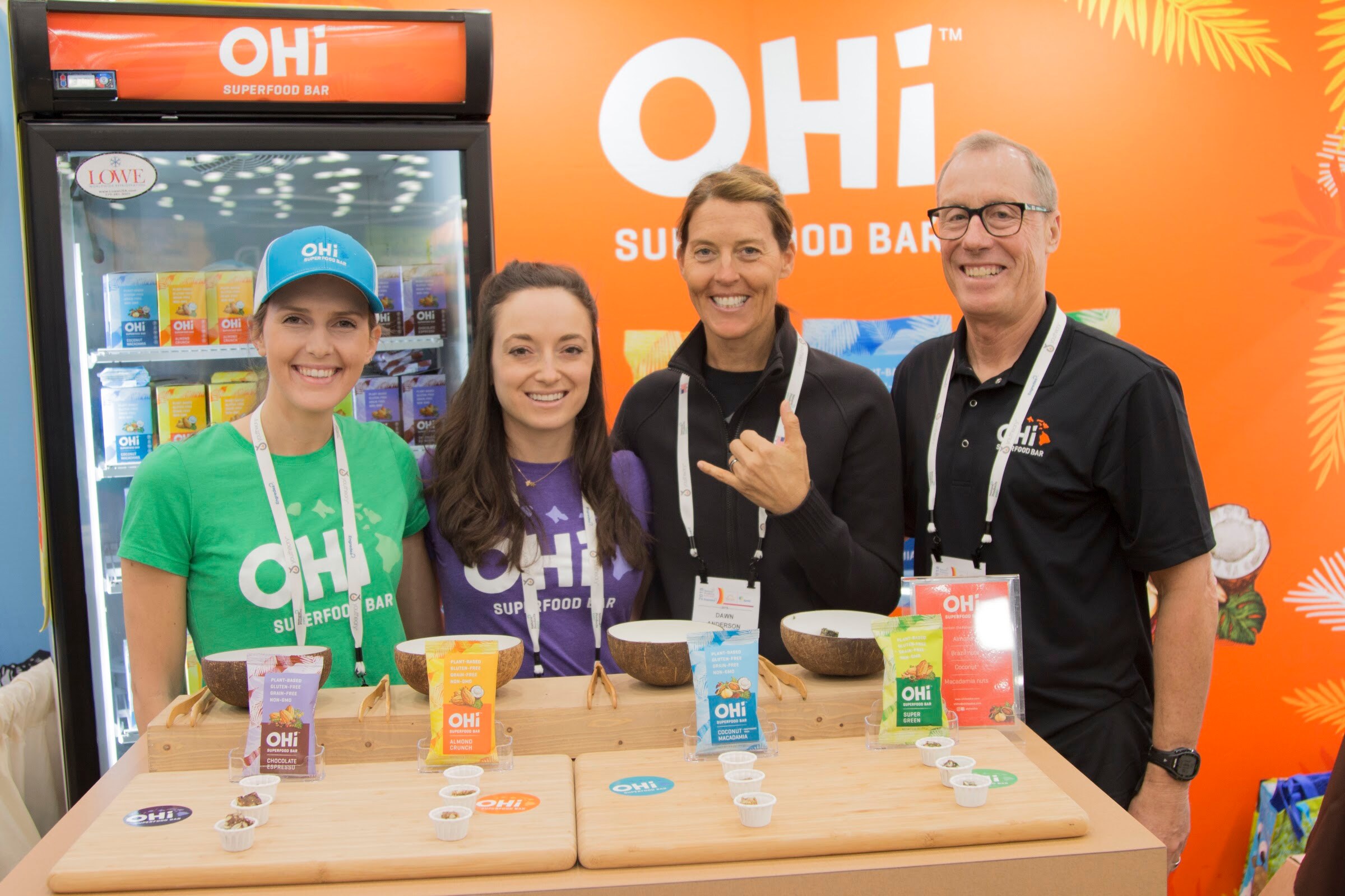 Four people in OHi branded shirts standing behind a wooden counter with coconut-shell sampling bowls in front of an orange OHi backdrop.