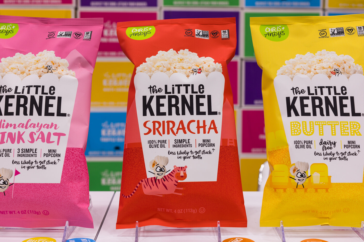 Three bags of The Little Kernel popcorn in pink, red and yellow in front of a colorful wall of boxes.