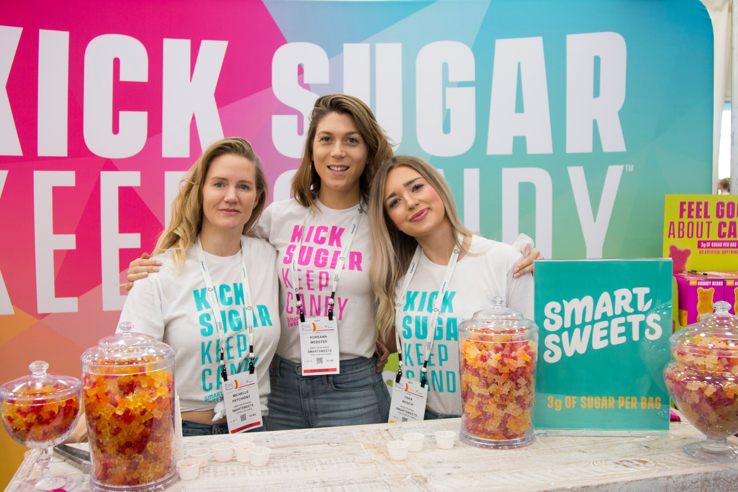 Three women in white 'Kick Sugar Keep Candy' t-shirts smiling behind a counter with glass jars of gummy candy and a Smart Sweets sign.