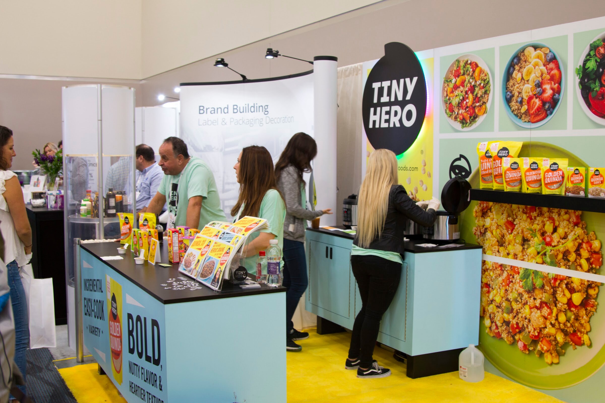 Booth with a large Tiny Hero logo, a yellow graphic of a grain bowl, and staff working at a light-blue cooking counter.