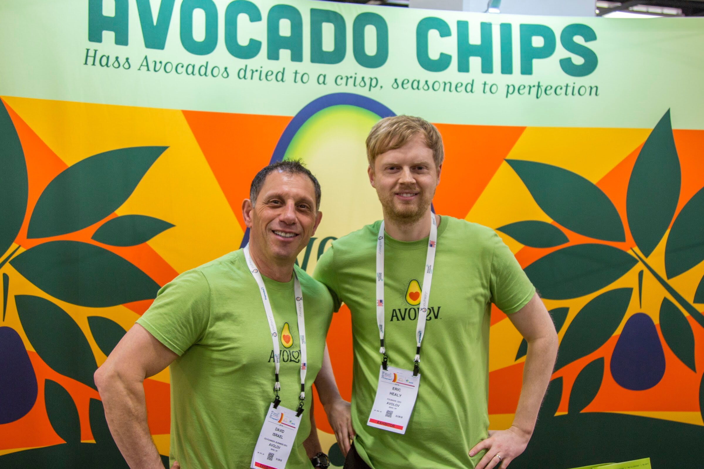 Two men with lanyards in matching green shirts posing in front of a colorful backdrop reading 'Avocado Chips'.