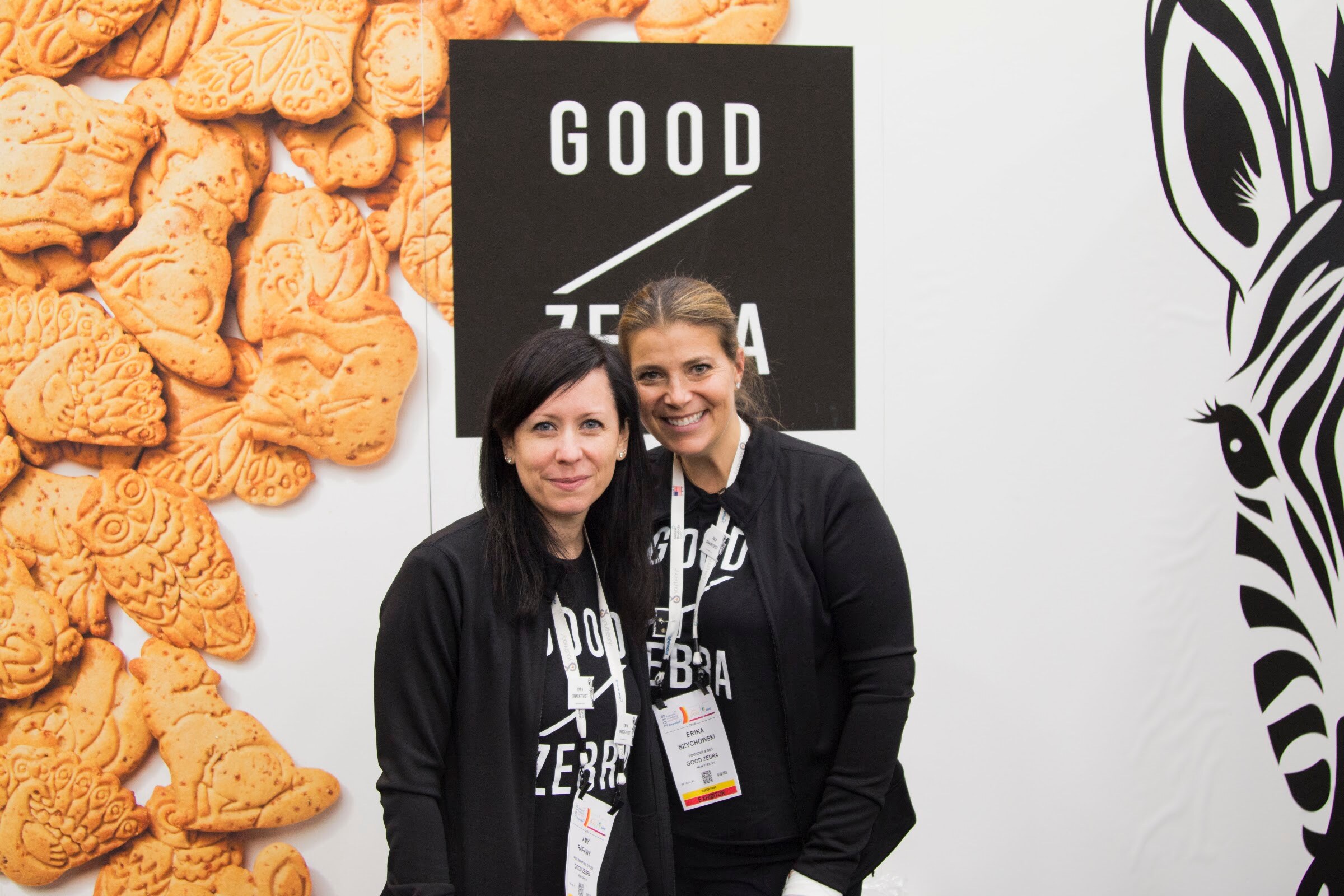 Two women in black shirts with a zebra logo standing in front of a wall printed with animal-shaped crackers and a Good Zebra sign.