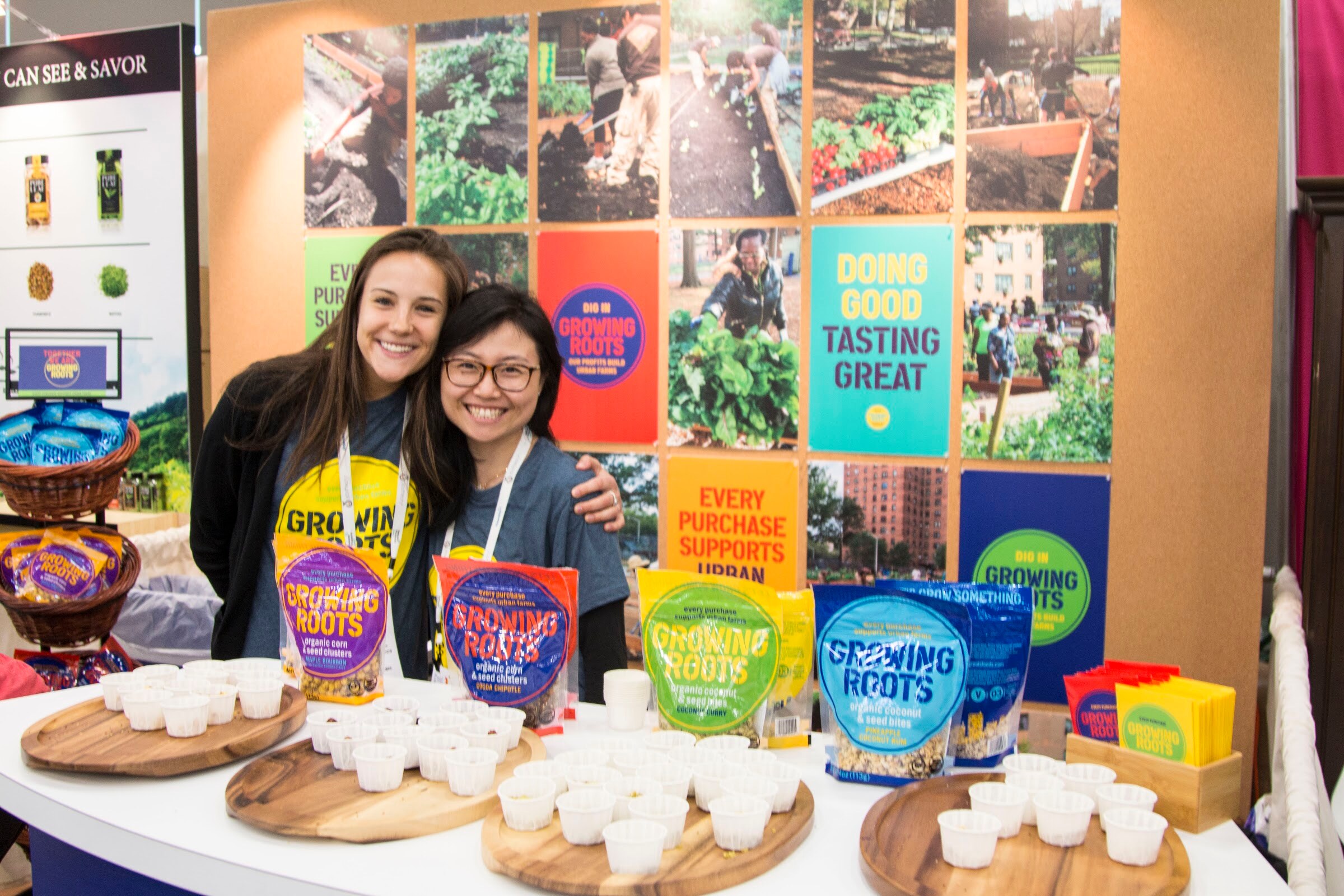 Two smiling women in grey shirts standing behind trays of sample cups and bags of Growing Roots snacks, with a wall of garden photos behind them.