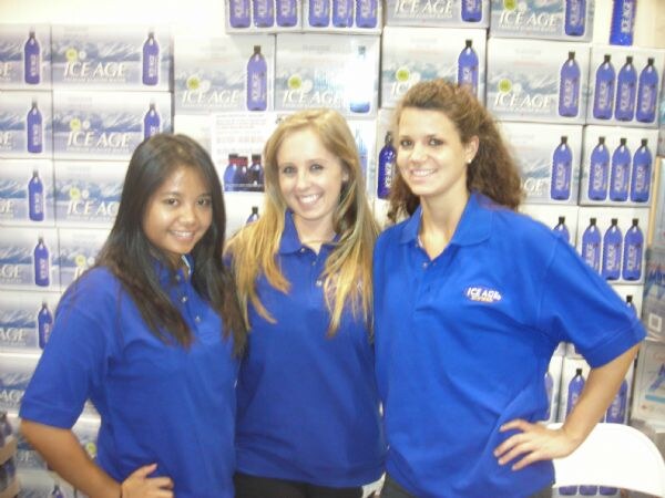 Three smiling women in blue polo shirts stand together in front of stacked cases of Ice Age water with purple-capped bottles; one shirt carries the Ice Age logo.