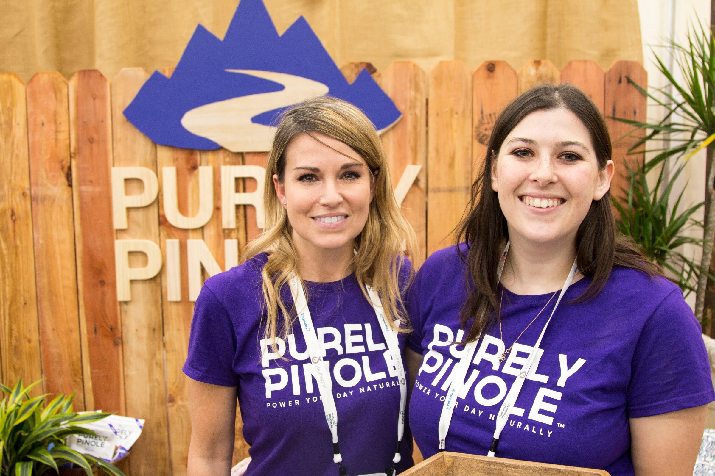 Two women in purple t-shirts with lanyards posing beneath a sign for Purely Pinole mounted on a wooden fence backdrop.