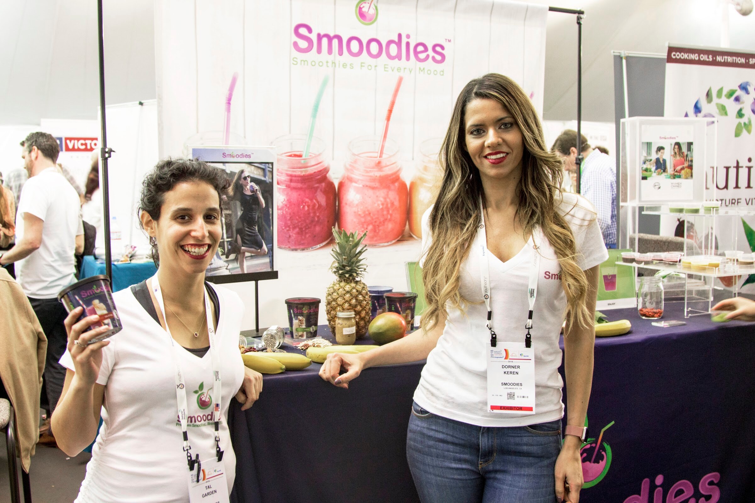 Two women in white Smoodies shirts smiling at a booth with jars of pink smoothies and a pineapple on the table.