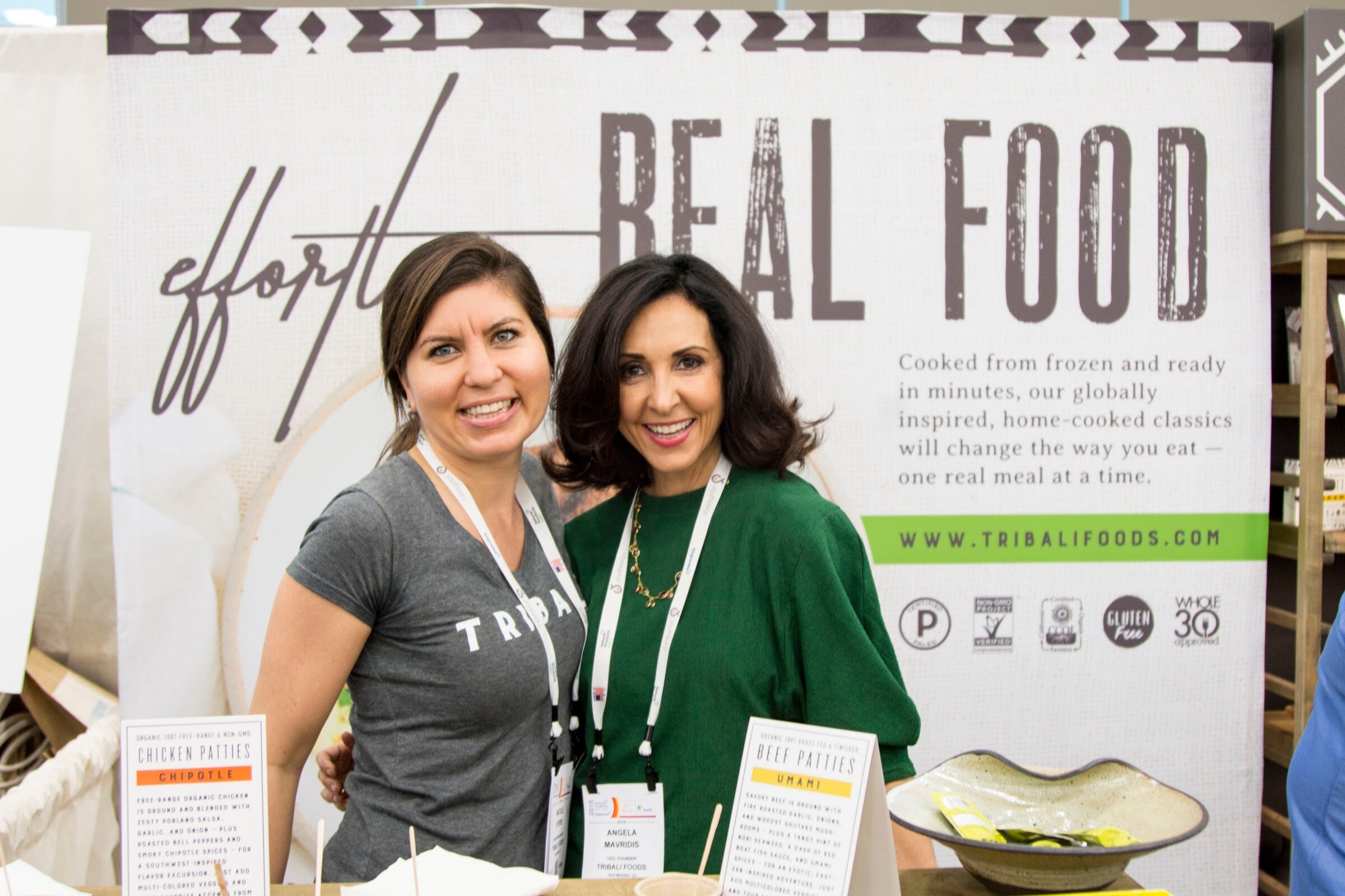 Two smiling women with lanyards standing in front of a Tribali Foods banner that reads 'effortless real food', with product cards on the counter.