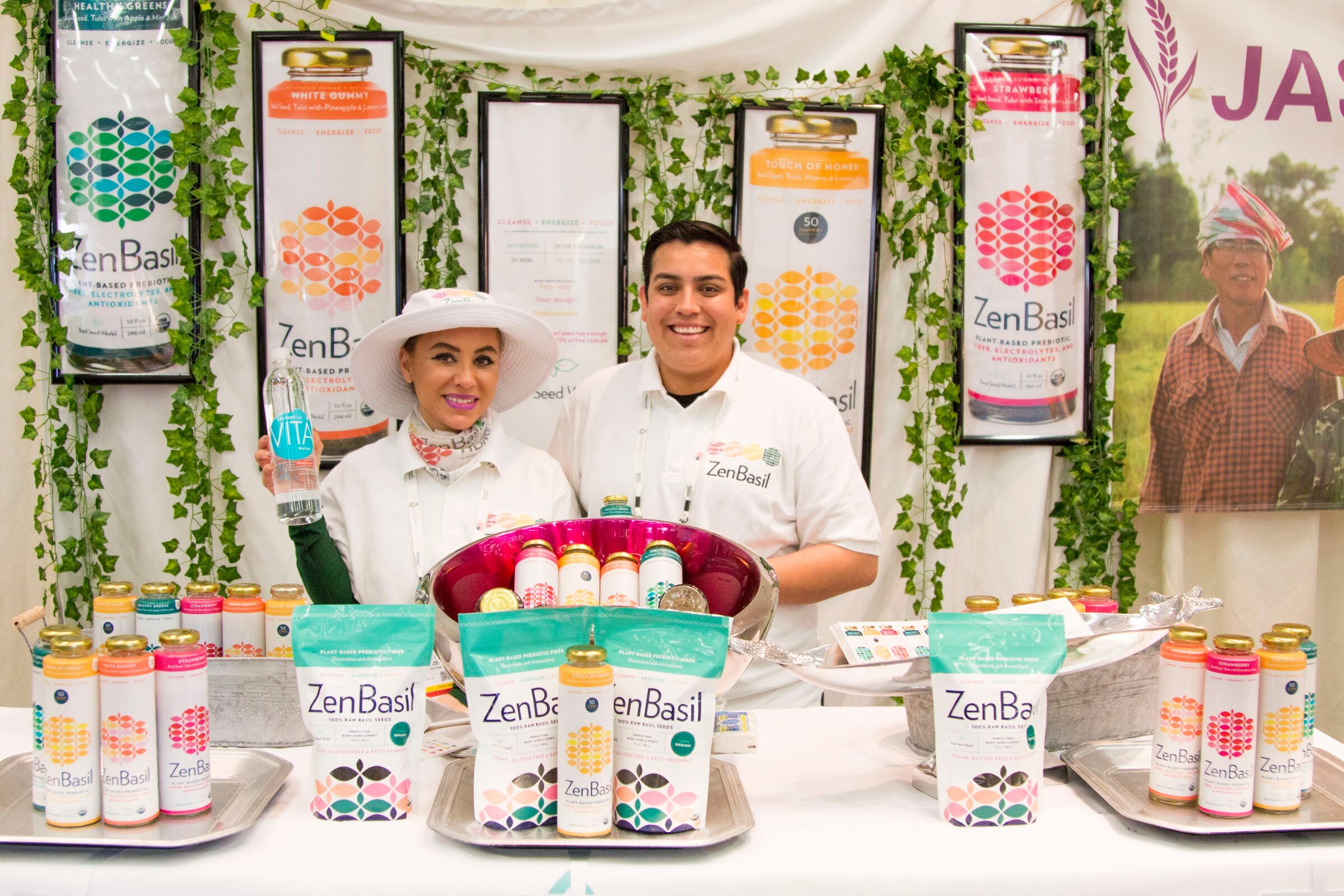 Woman in a wide-brim white hat and a man in white standing behind a table of ZenBasil bottles and bags with vines hanging on the wall behind.