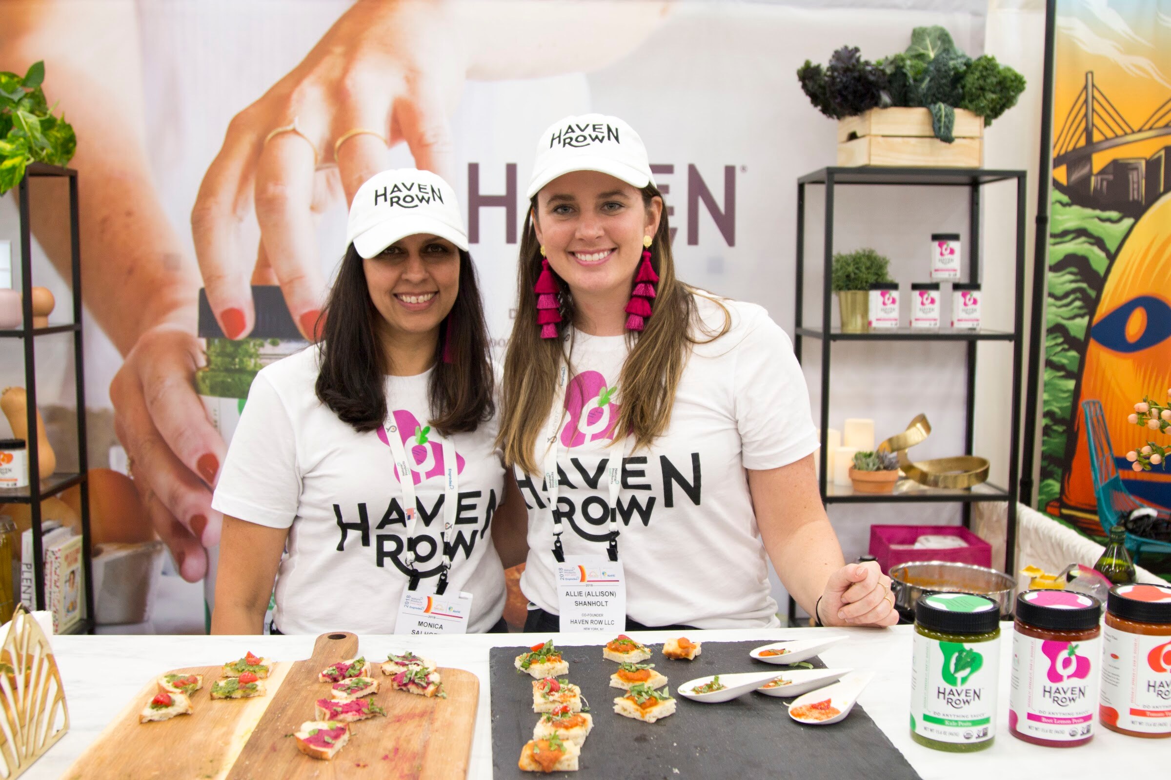 Two smiling women in white Haven Row shirts and caps behind a counter with plates of appetizers and jars of sauce.