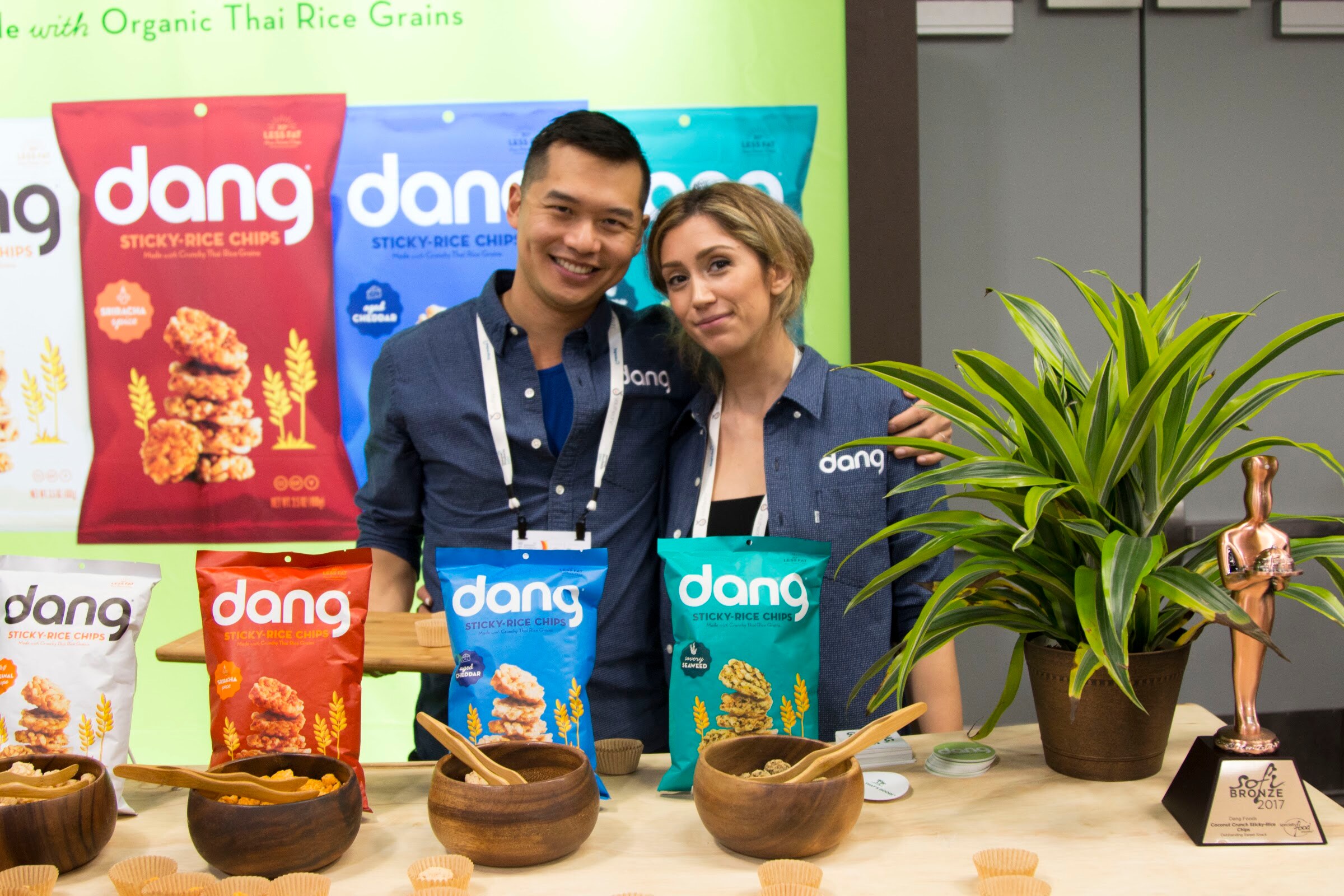 Man and woman in dark Dang shirts posing behind wooden bowls of chips, bags of Dang sticky-rice chips and a bronze award statuette.