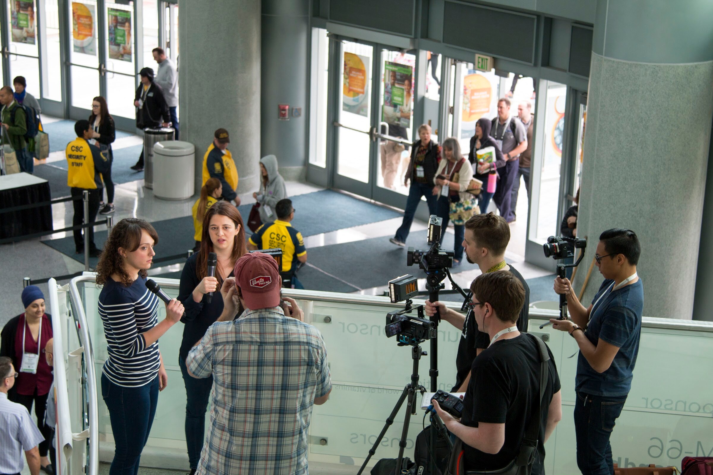 High angle of a lobby where reporters hold microphones and camera operators on tripods film, with attendees entering through glass doors.