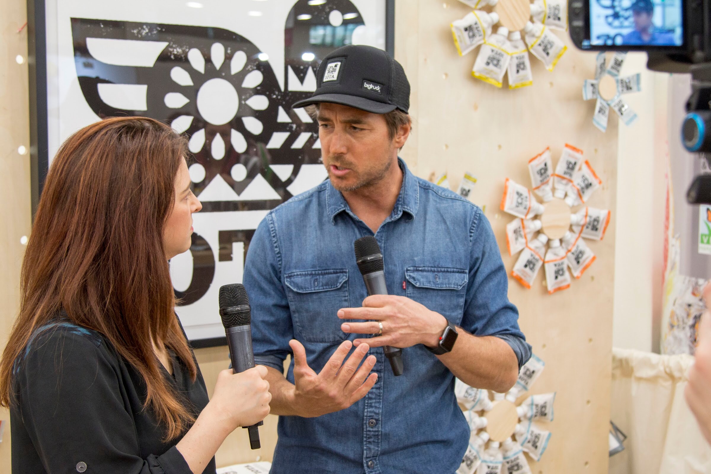 Woman with a microphone and man in a baseball cap and denim shirt talking in front of a wooden wall with packaged snacks, with a camera at the right.