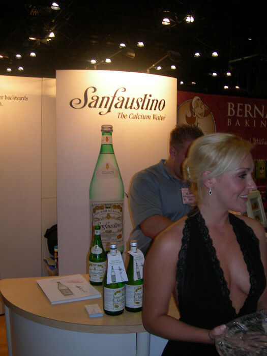 A blonde woman in a black halter top stands at a booth with a Sanfaustino The Calcium Water display and green bottles on the counter, while a man in a gray shirt stands behind.