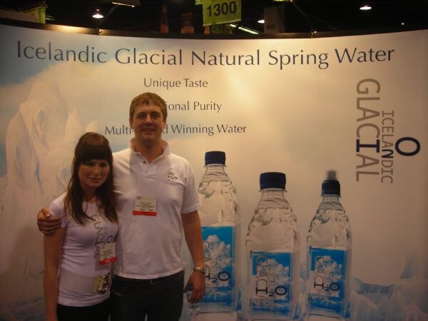 A man in a white polo with his arm around a woman in a gray top stands before an Icelandic Glacial Natural Spring Water backdrop showing three large water bottles.