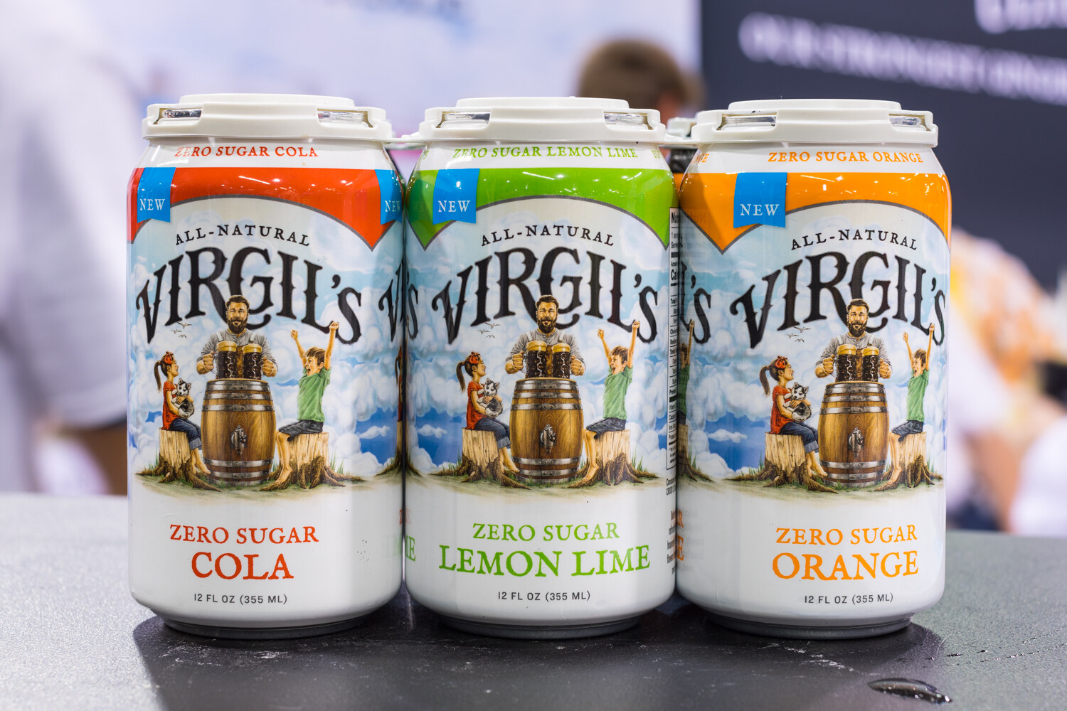 Three Virgil's zero sugar soda cans in cola, lemon lime and orange with a blurred show floor behind.