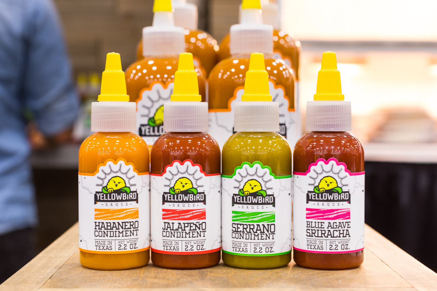 Row of four yellow-topped Yellowbird sauce bottles on a wooden riser, with more bottles behind.