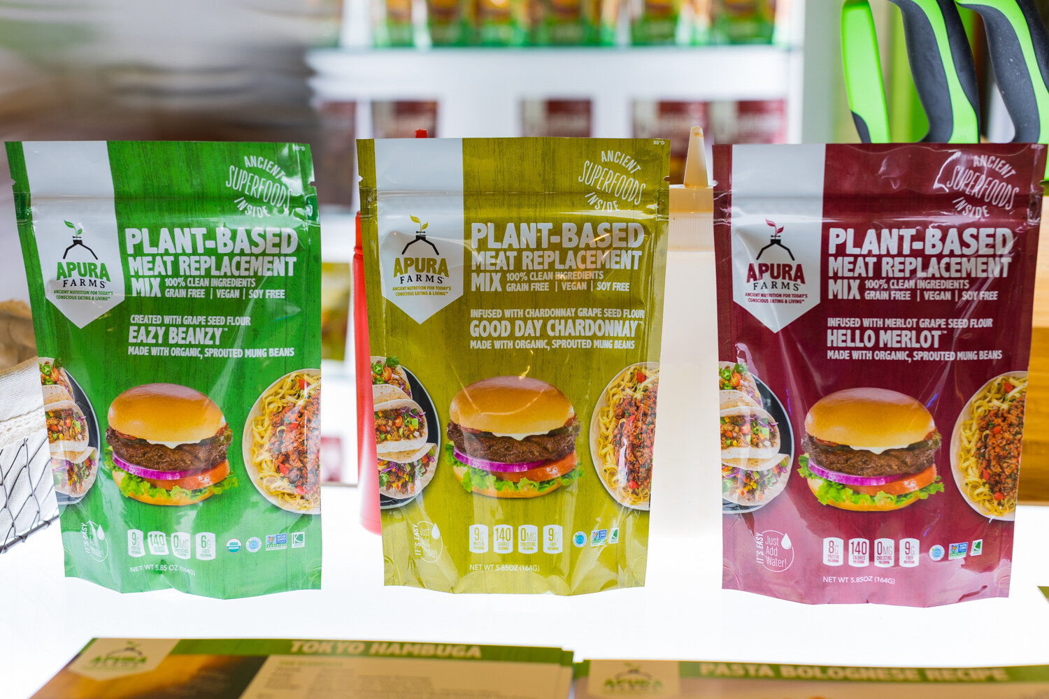 Three green, olive and burgundy Apura Farms pouches on a lit white shelf with recipe cards below.