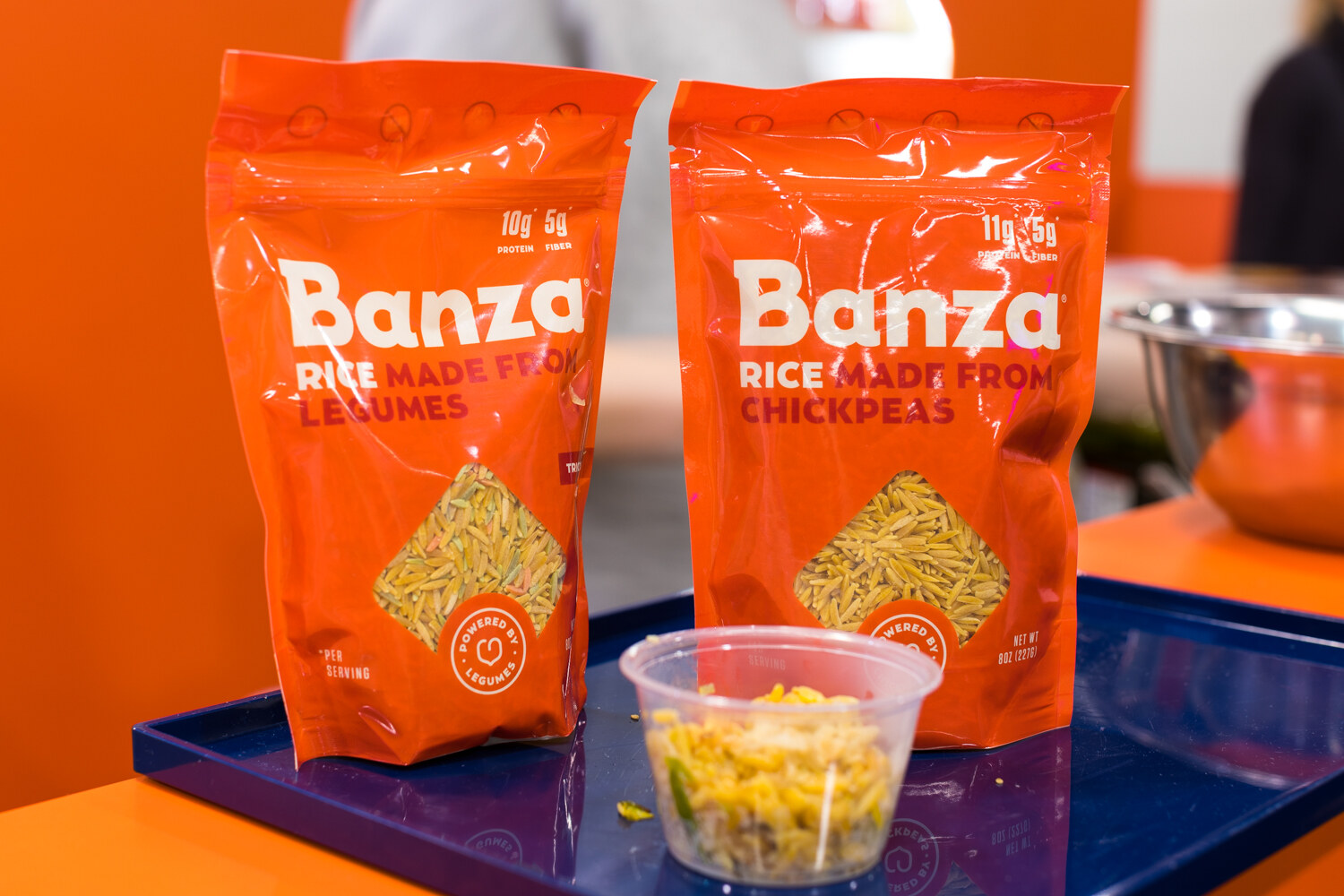 Two orange Banza bags on a navy tray with a clear sample cup of cooked rice, against an orange wall.