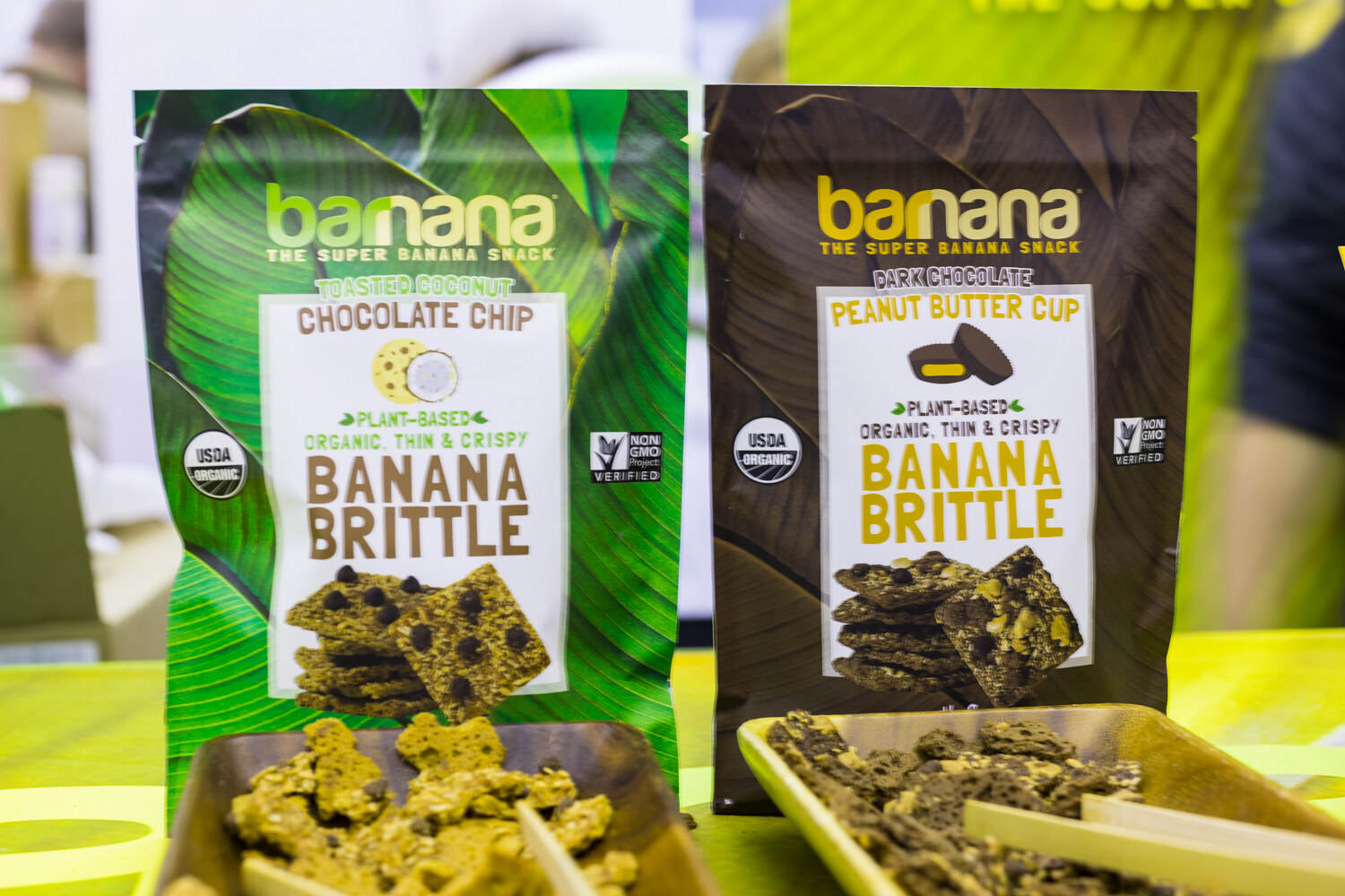 Two Barnana Banana Brittle bags in green and brown with wooden trays of brittle pieces in front on a lime green table.
