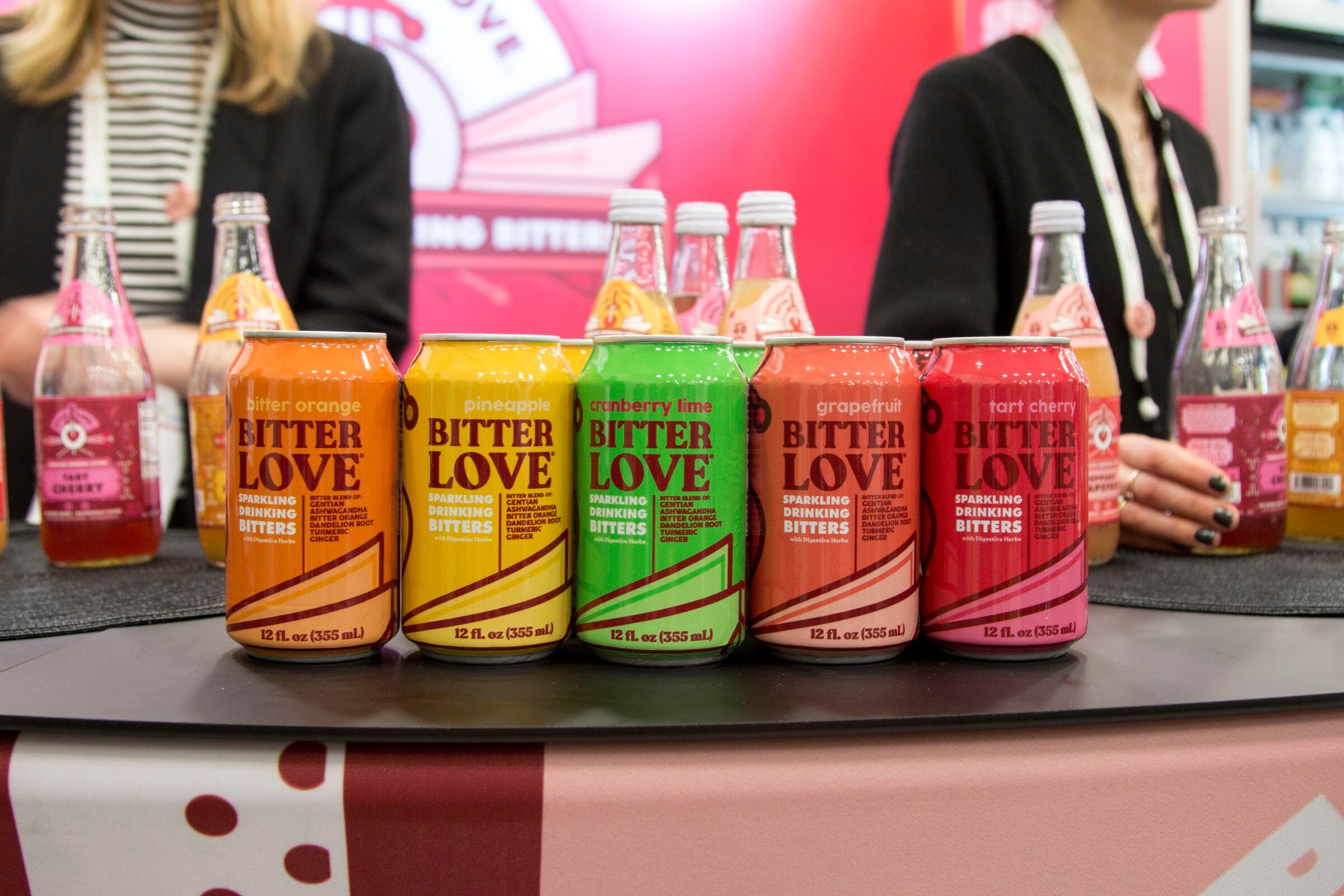 Five Bitter Love cans in orange, yellow, green, coral and red with glass bottles and two staffers' torsos behind them.