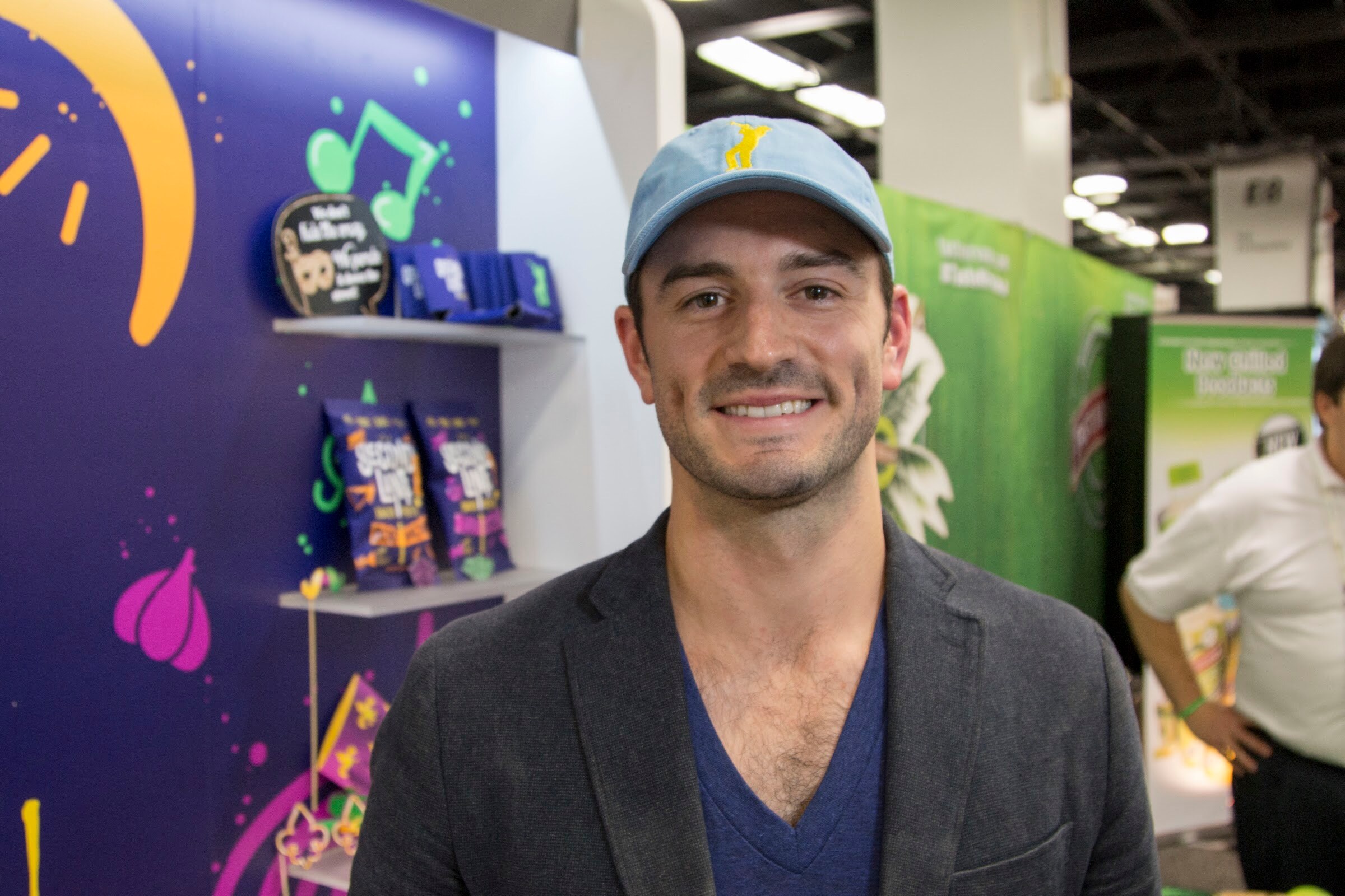 A man in a light blue baseball cap smiling in front of a purple wall with shelves of snack bags.