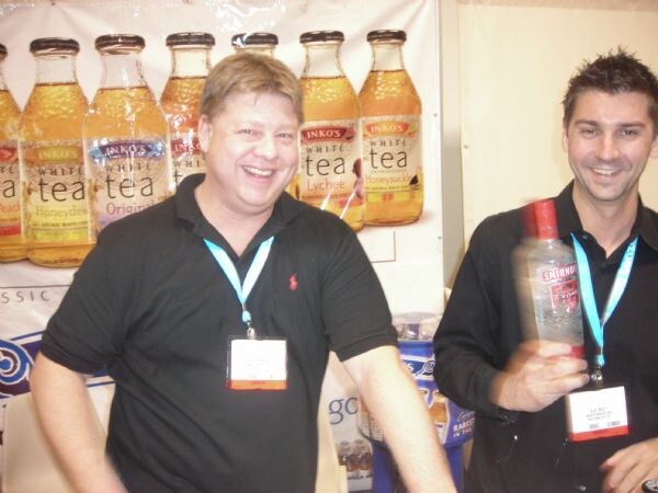 Two smiling men wearing badges and lanyards stand in front of a display of Inko's White Tea bottles; the man at right holds a Smirnoff bottle.