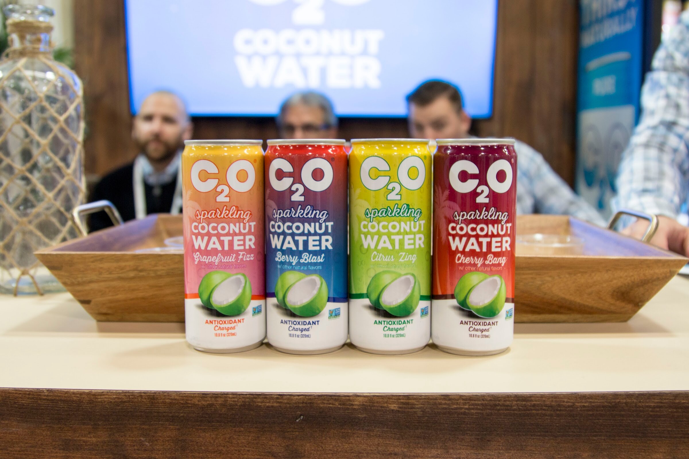 Four colorful C2O cans in a row on a tray with three men seated behind and a coconut water screen above.